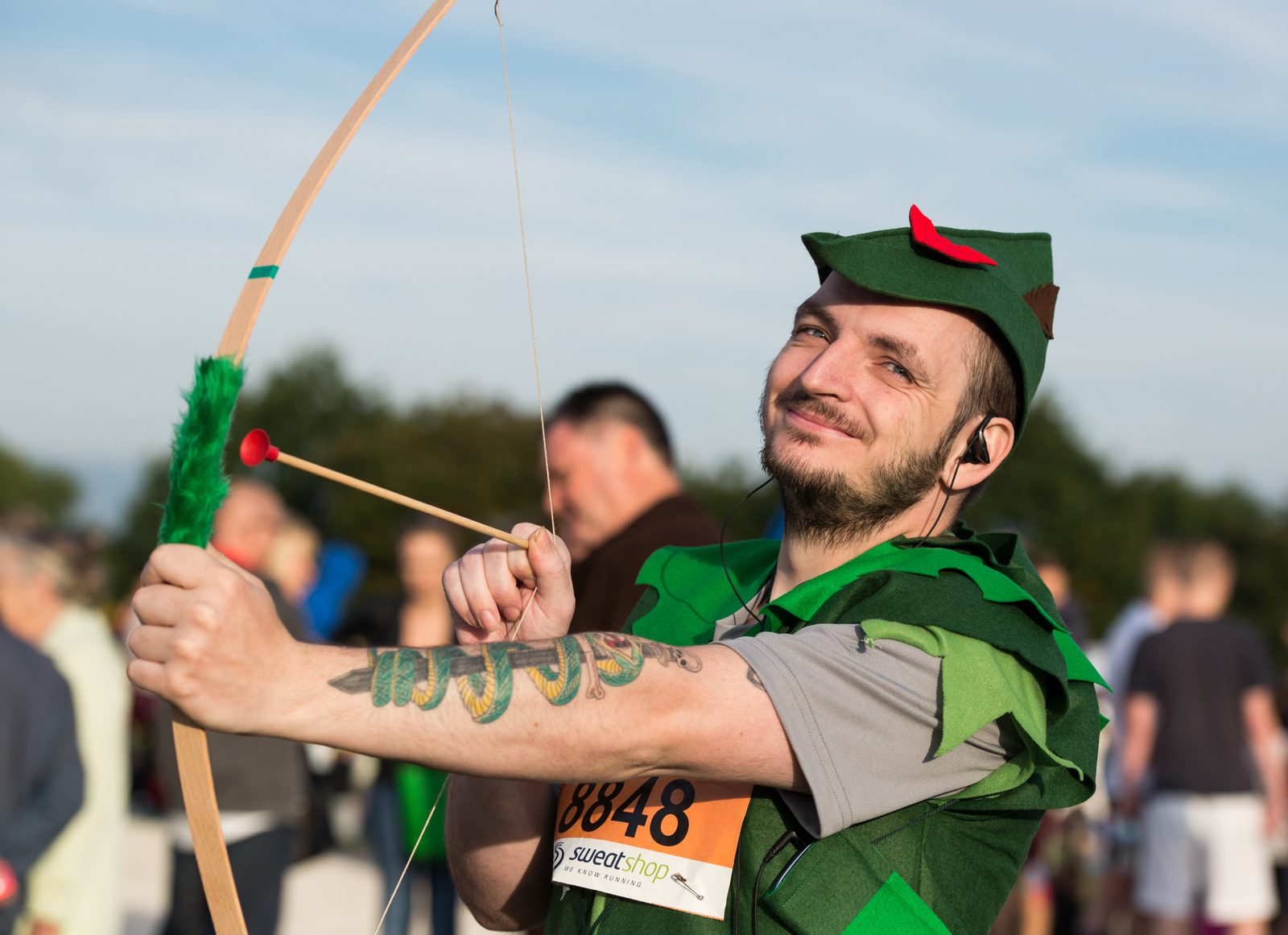 A person dressed in a green, Robin Hood-style costume holds a toy bow and arrow, aiming it playfully. The individual is smiling and has visible tattoos on their forearm. They are outdoors with a crowd of people in the background.