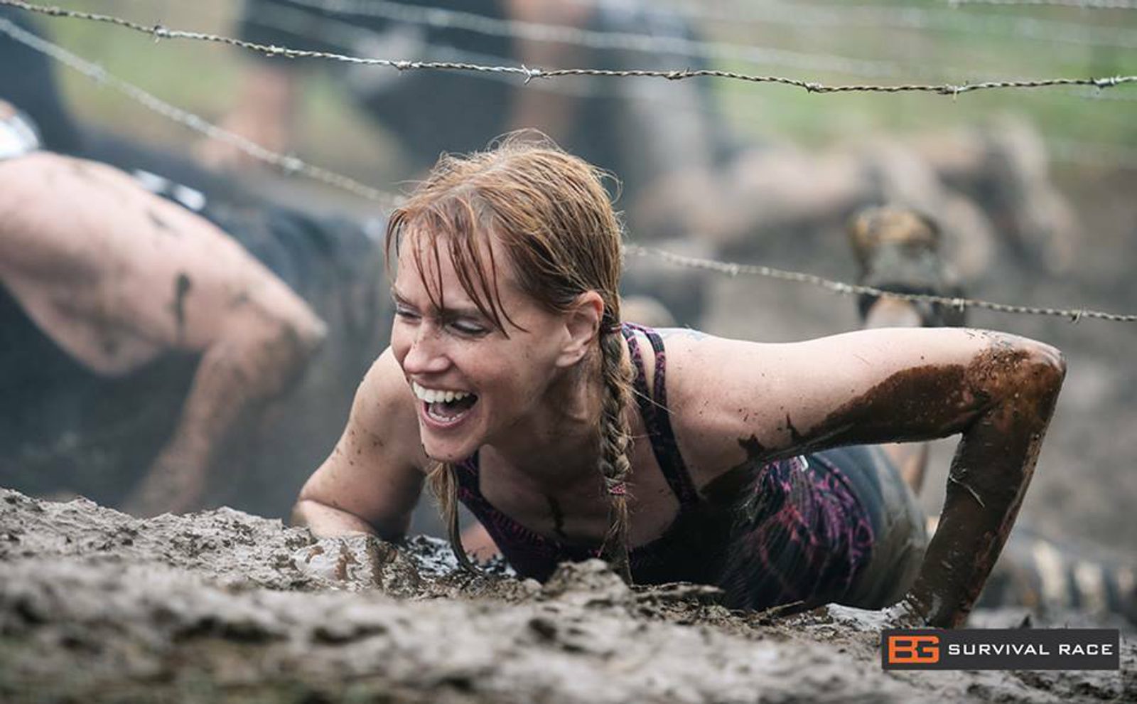 A woman with braided hair, covered in mud, is smiling as she crawls under barbed wire during an obstacle race. The event logo "BG Survival Race" is visible in the bottom right corner. Other participants are also crawling through the muddy terrain in the background.