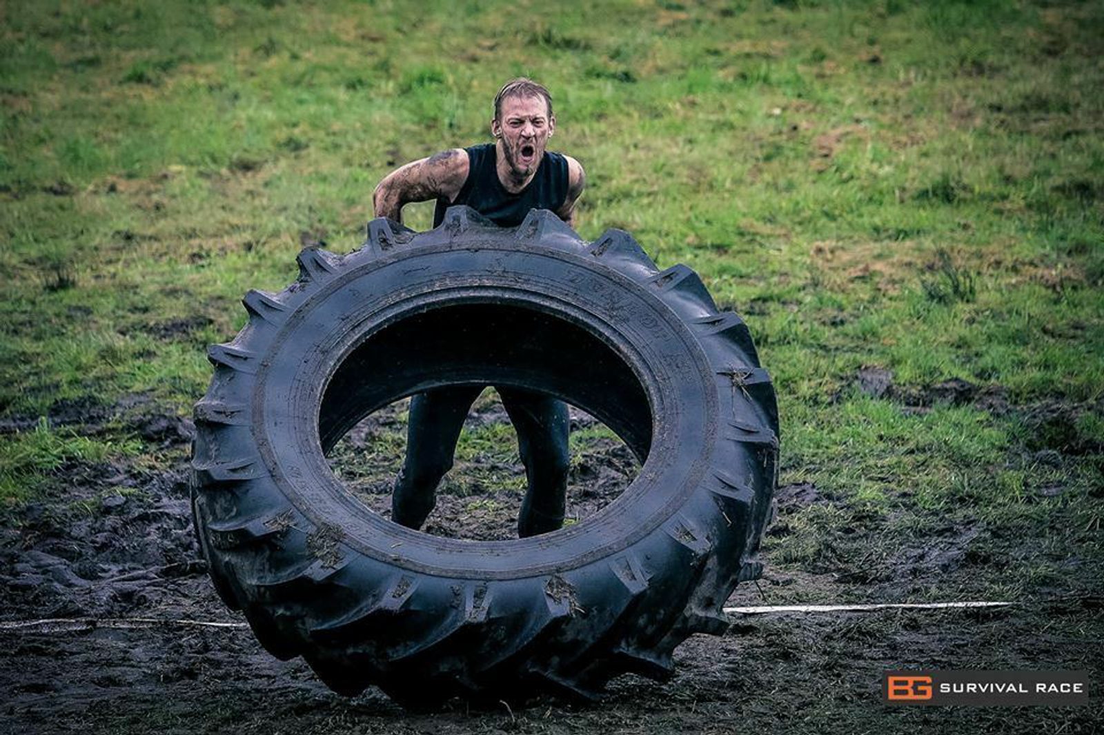 A person in athletic gear, covered in mud, exerts effort while lifting a large tractor tire during an outdoor obstacle course race. The backdrop features a grassy, muddy area with the event logo in the bottom right corner.