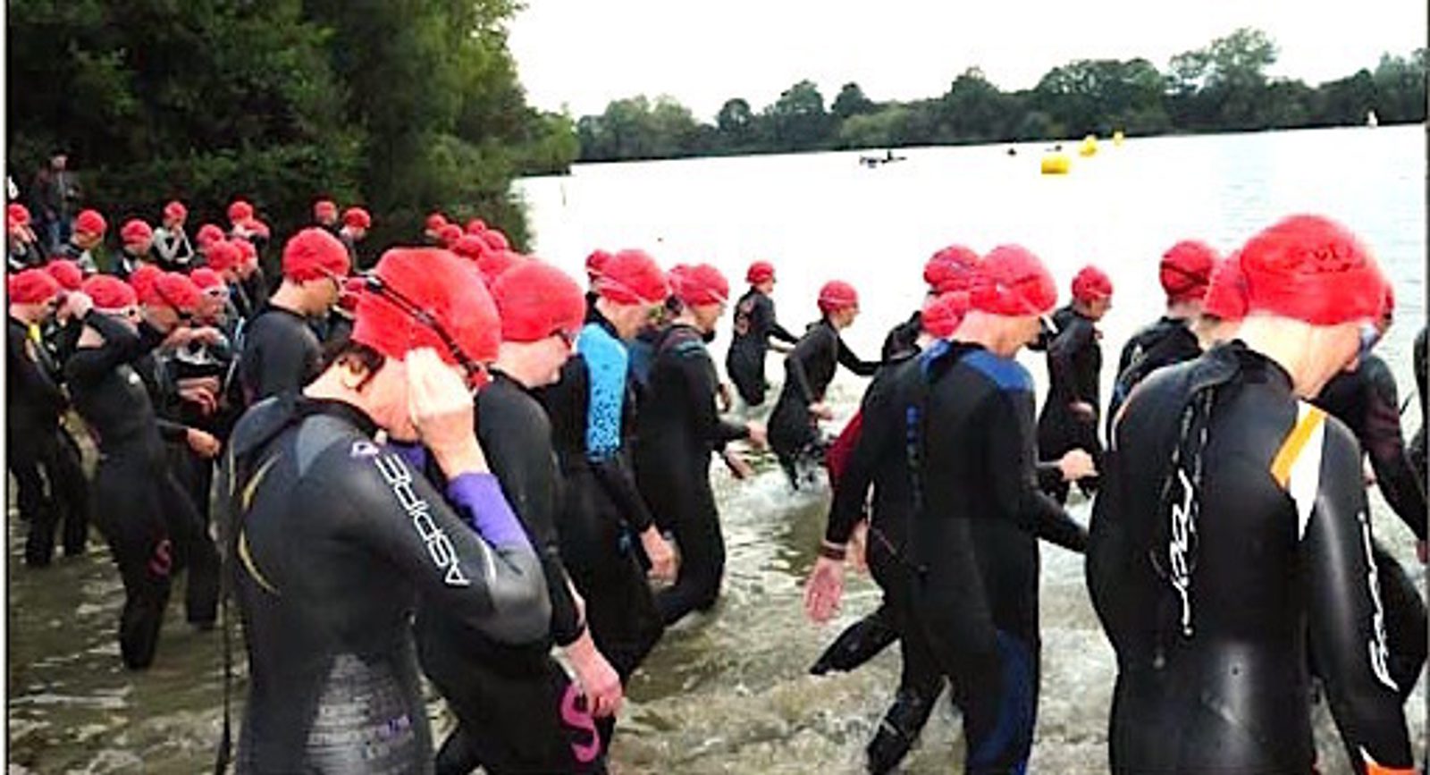 Swimmers in wetsuits and red swim caps are entering the water from a lakeshore, preparing for a race. The background shows trees, buoys in the water, and a cloudy sky.