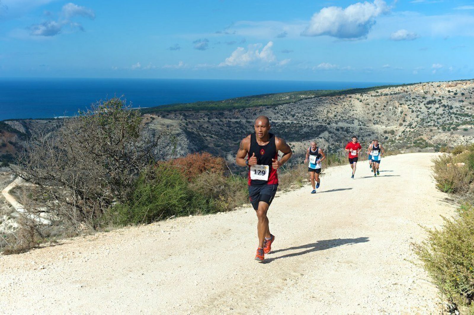 Four runners are participating in a race on a dirt road winding through a hilly landscape with sparse vegetation. The blue ocean and clear sky form the scenic backdrop. The leading runner wears a black and red outfit, with a readable bib number, 139.