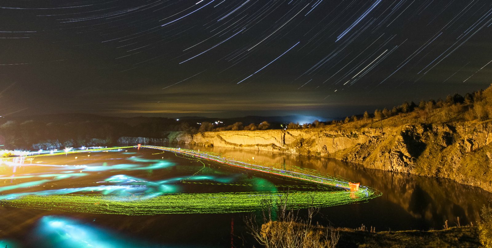 A long-exposure nighttime photograph of a lake surrounded by rugged terrain and trees. Star trails form arcs in the sky, and colorful, wavy light trails in green, red, and yellow float over the water near the shore, creating a vibrant, surreal scene.