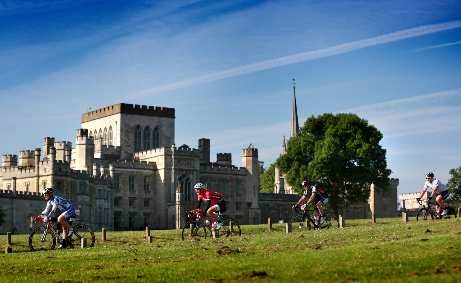 Four cyclists ride on a grassy path with a large historic castle and a church spire in the background. The sky is blue with wispy clouds, and the terrain appears slightly hilly, surrounded by greenery.