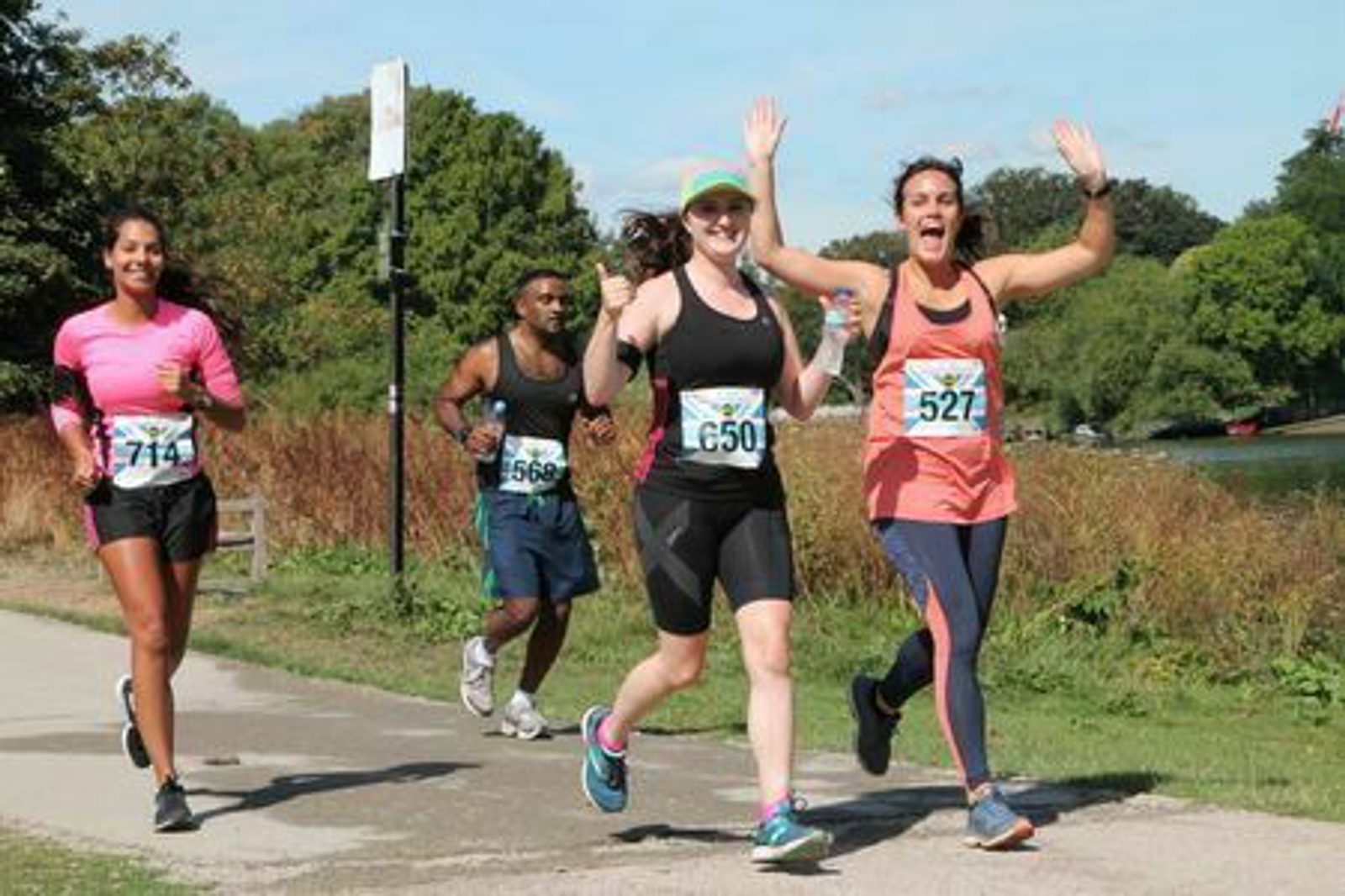 Four runners, two women and two men, are participating in a race along a scenic path with trees and a body of water in the background. The runners are smiling and waving, wearing race bibs, athletic gear, and running shoes. The woman in pink looks particularly joyful.