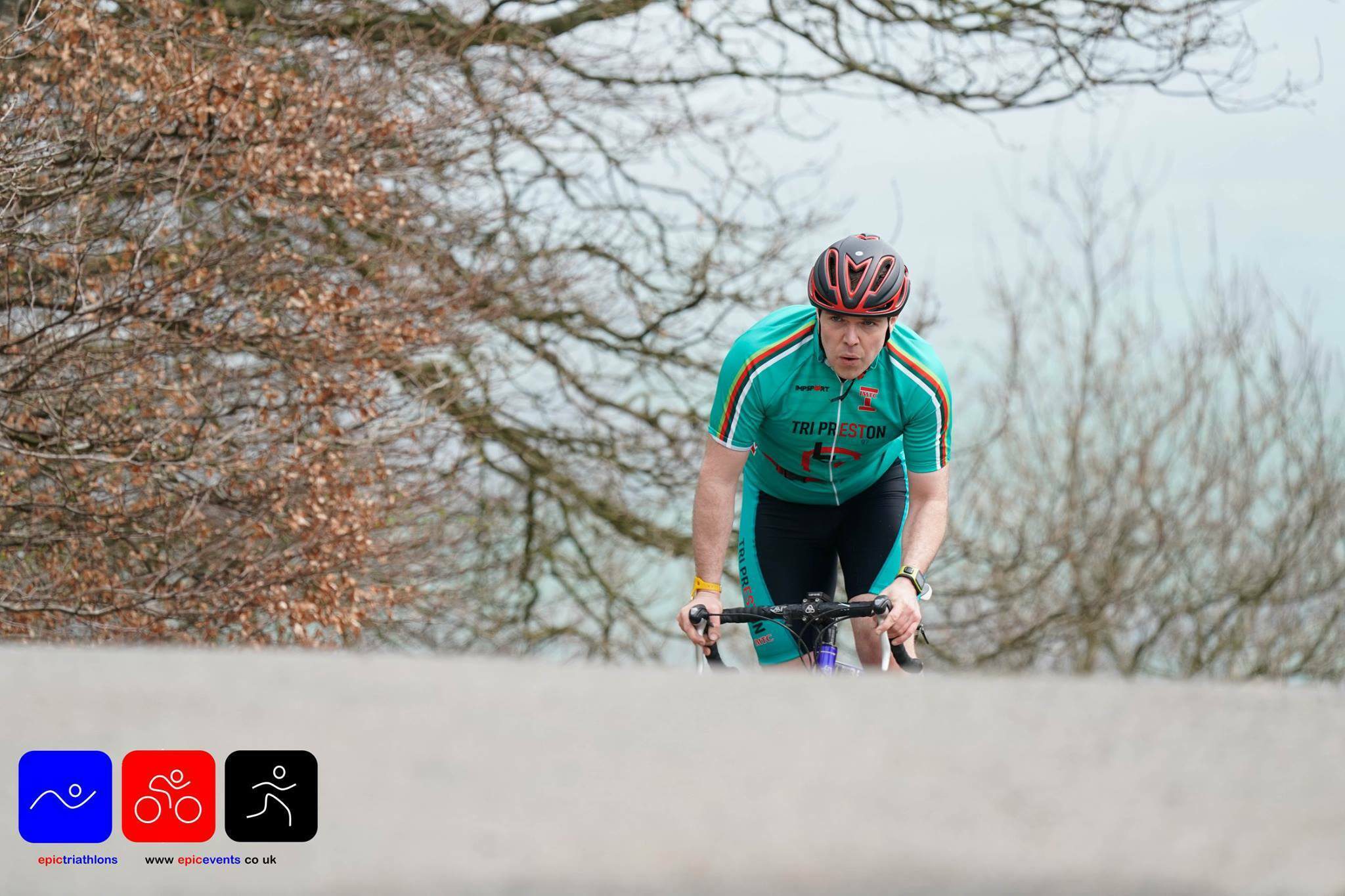 A cyclist wearing a green and red jersey and black helmet rides up a hill with bare trees in the background. There are three logos at the bottom left corner of the image, indicating the website and nature of the triathlon event.