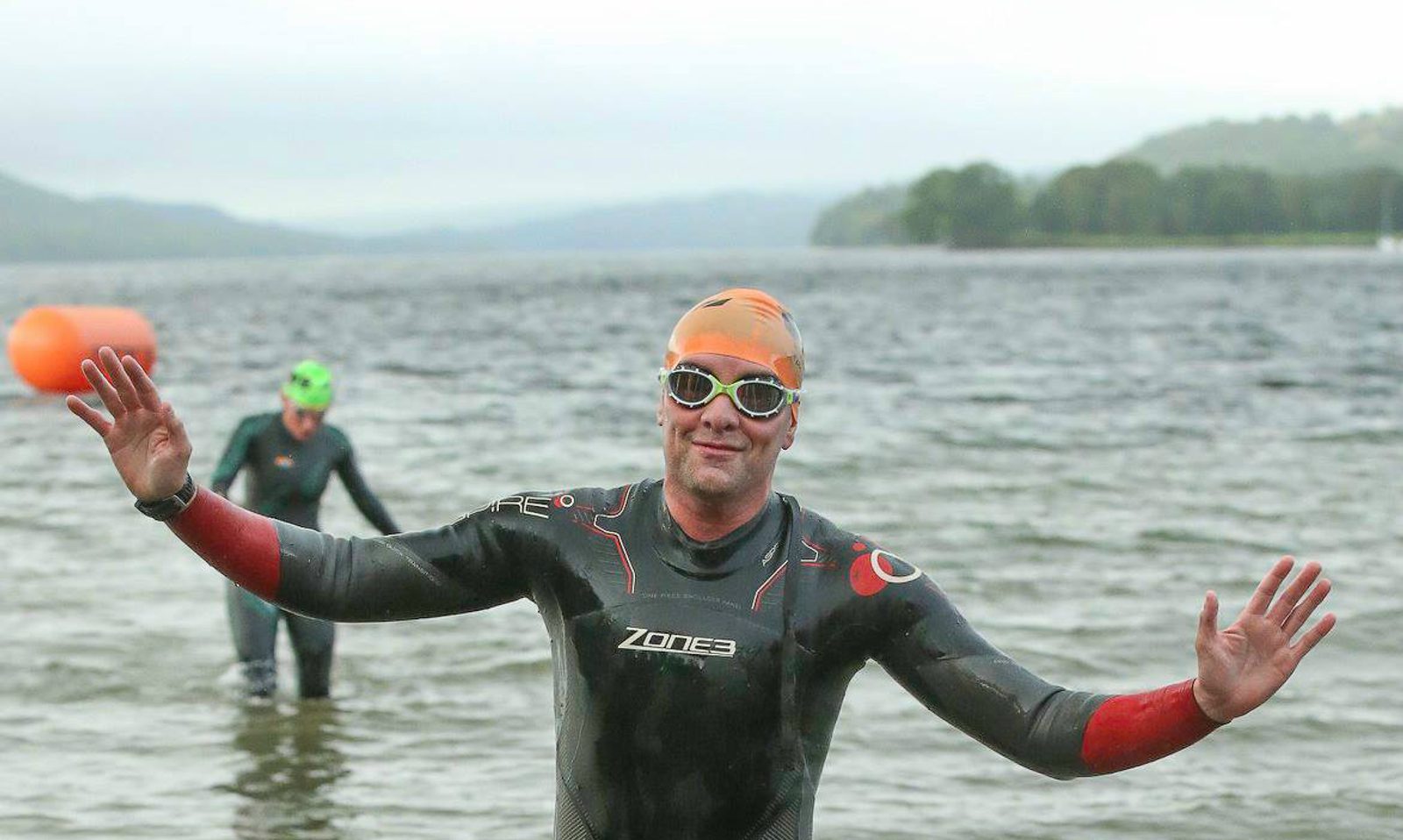 A person wearing a black wetsuit, orange swim cap, and white goggles smiles and raises their arms while standing in shallow water. Another swimmer in a green swim cap is in the background. The scene is set against a calm lake with a distant tree-lined shore.