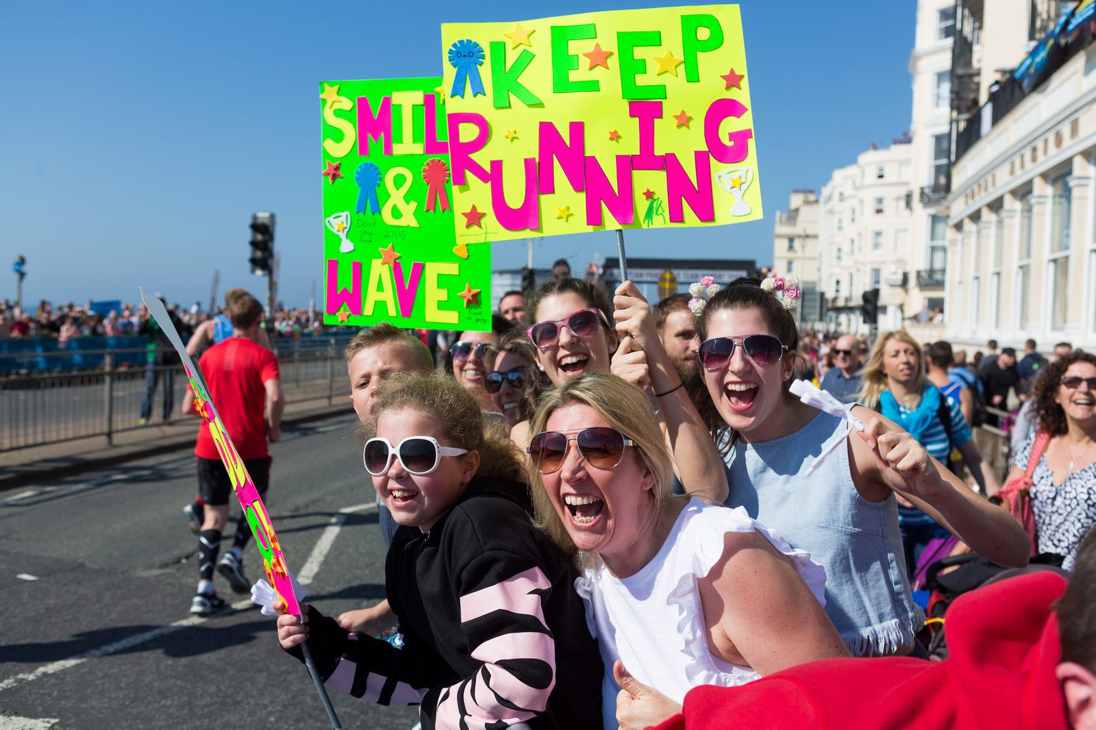 A group of enthusiastic people, some holding a vibrant neon sign with decorations, cheer and smile at a race event. The sign reads "KEEP SMILING & RUNNING" with colorful letters and symbols. The sunny day adds vibrance to the lively atmosphere.