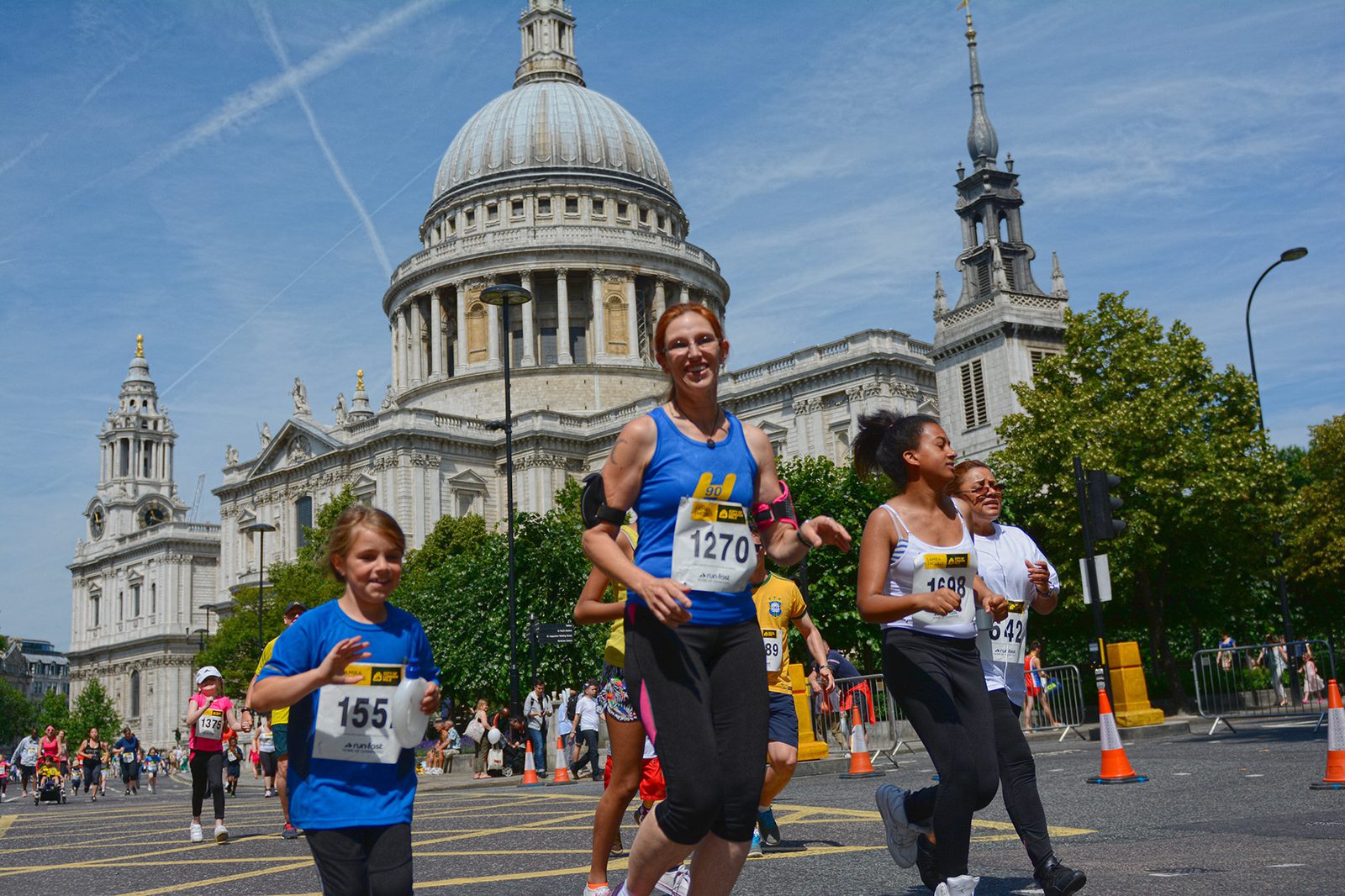 A group of smiling runners, including adults and a child, participate in a race in front of the majestic St. Paul's Cathedral on a sunny day. The runners wear numbered bibs and athletic attire, with onlookers and lush, green trees in the background.