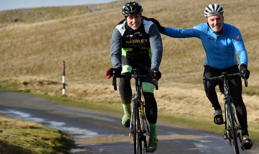 Two cyclists are riding on a country road with grassy hills in the background. One cyclist in a dark outfit has his arm around the shoulder of another cyclist in a blue jacket, both smiling and enjoying the ride. The weather appears to be cool and dry.