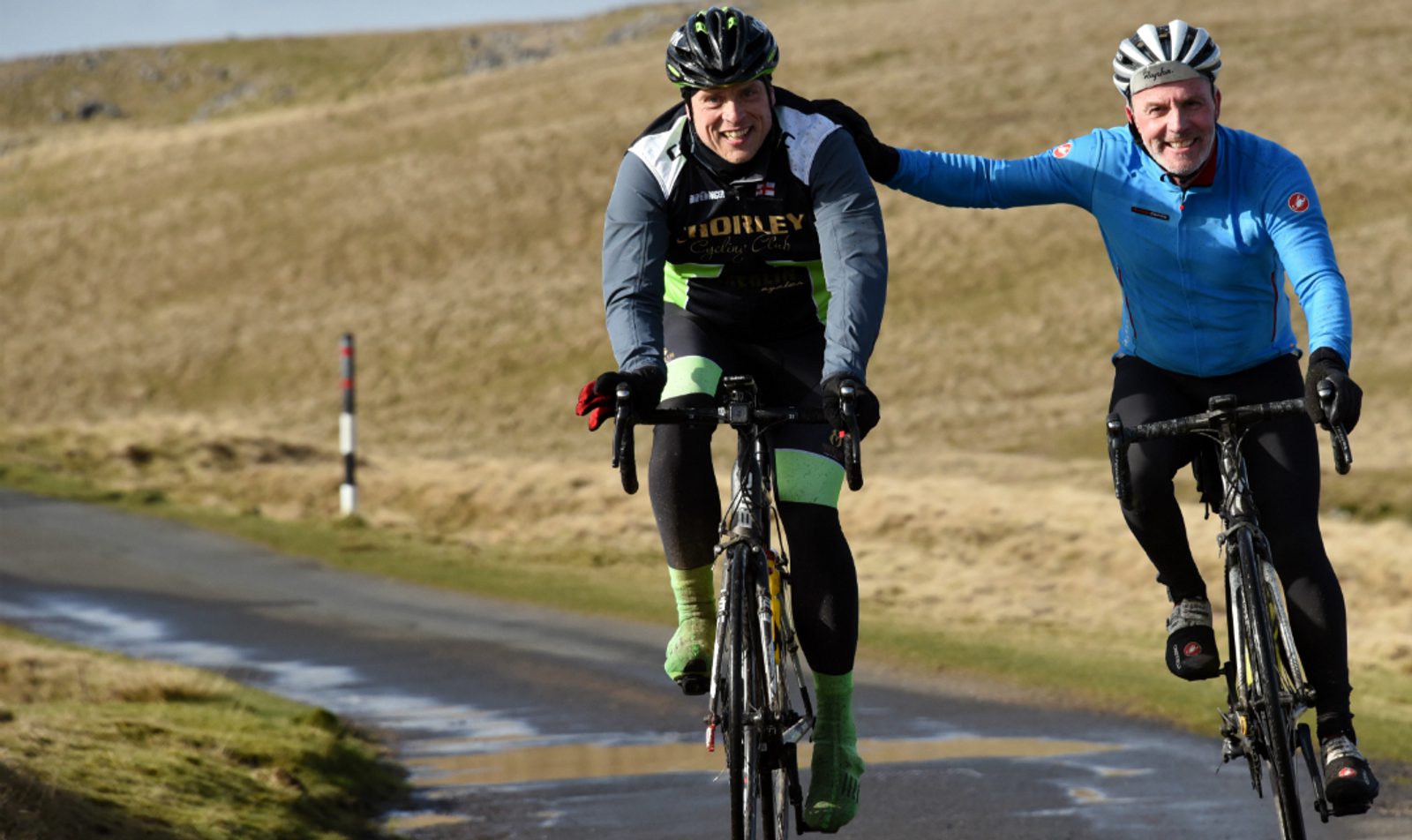 Two cyclists are riding on a country road with grassy hills in the background. One cyclist in a dark outfit has his arm around the shoulder of another cyclist in a blue jacket, both smiling and enjoying the ride. The weather appears to be cool and dry.