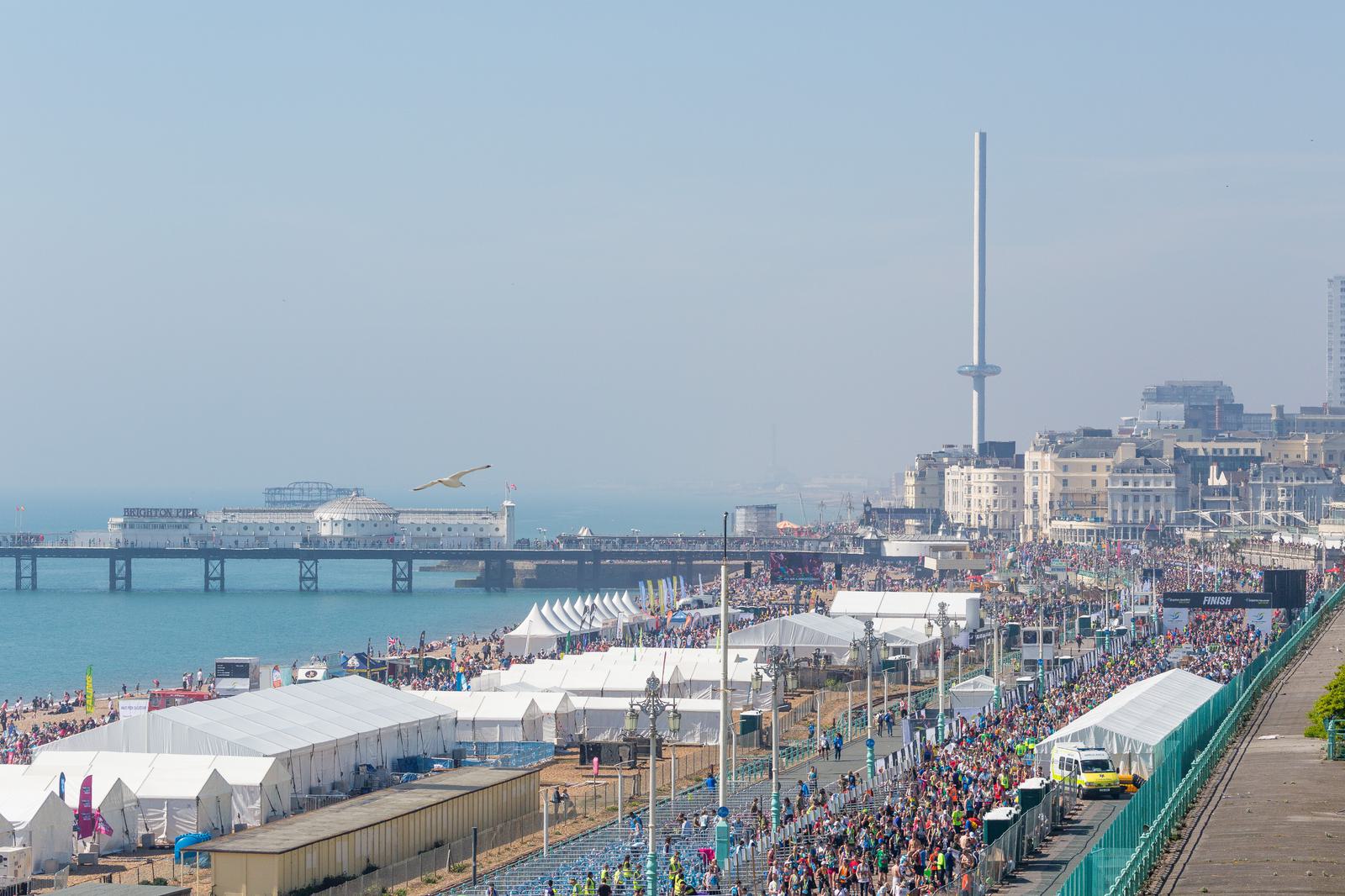An aerial view of a bustling seaside promenade, lined with white tents and filled with crowds of people. The pier extends into the sea on the left, while tall, modern structures rise in the background. A bird flies in the clear sky above the scene.