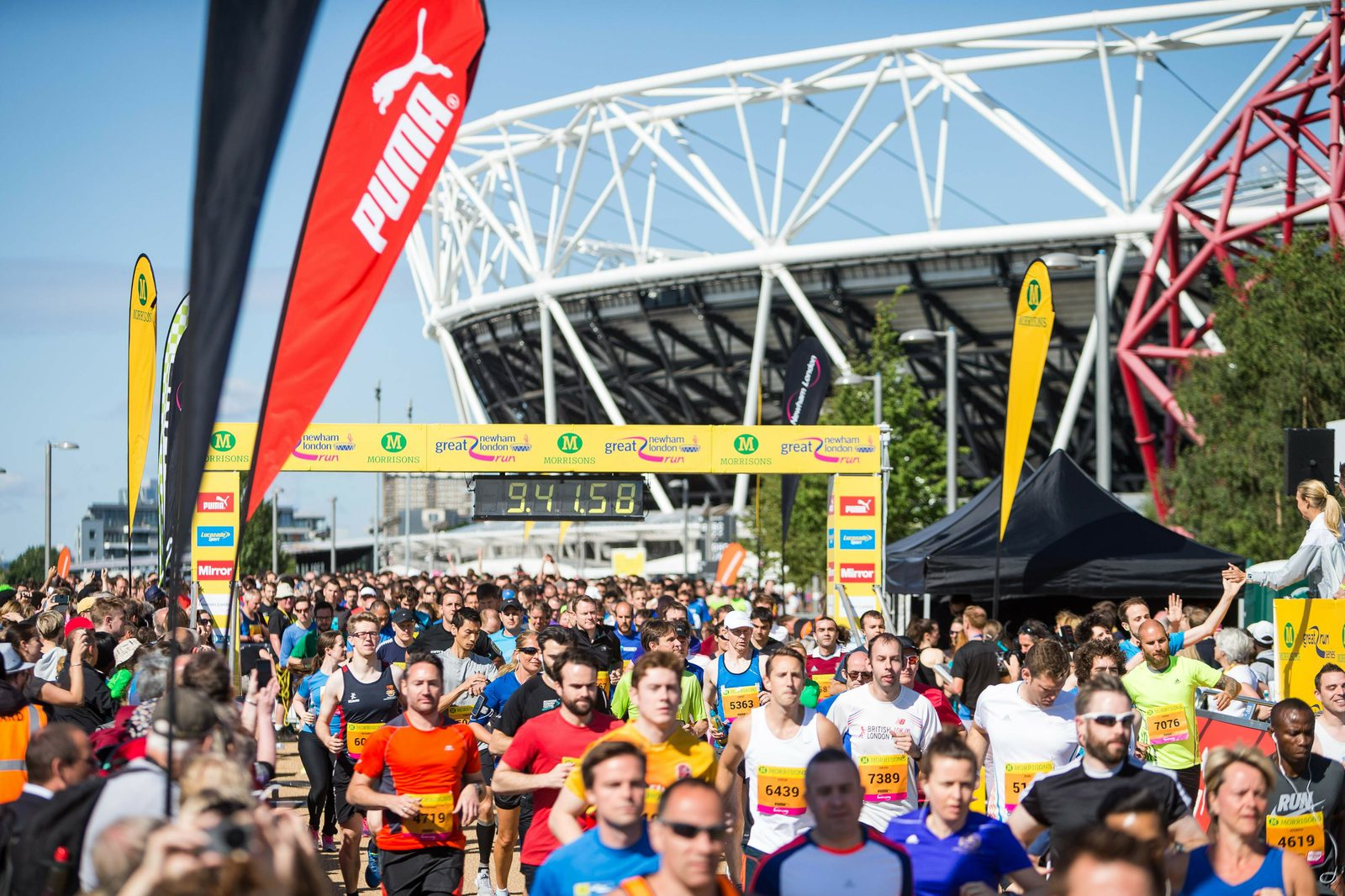 A large group of runners, wearing colorful athletic gear, participating in a race near a stadium. The runners are seen passing under a yellow finish line banner, with timing clocks showing 9:41:58. Bright red and green sponsor flags are visible in the foreground.