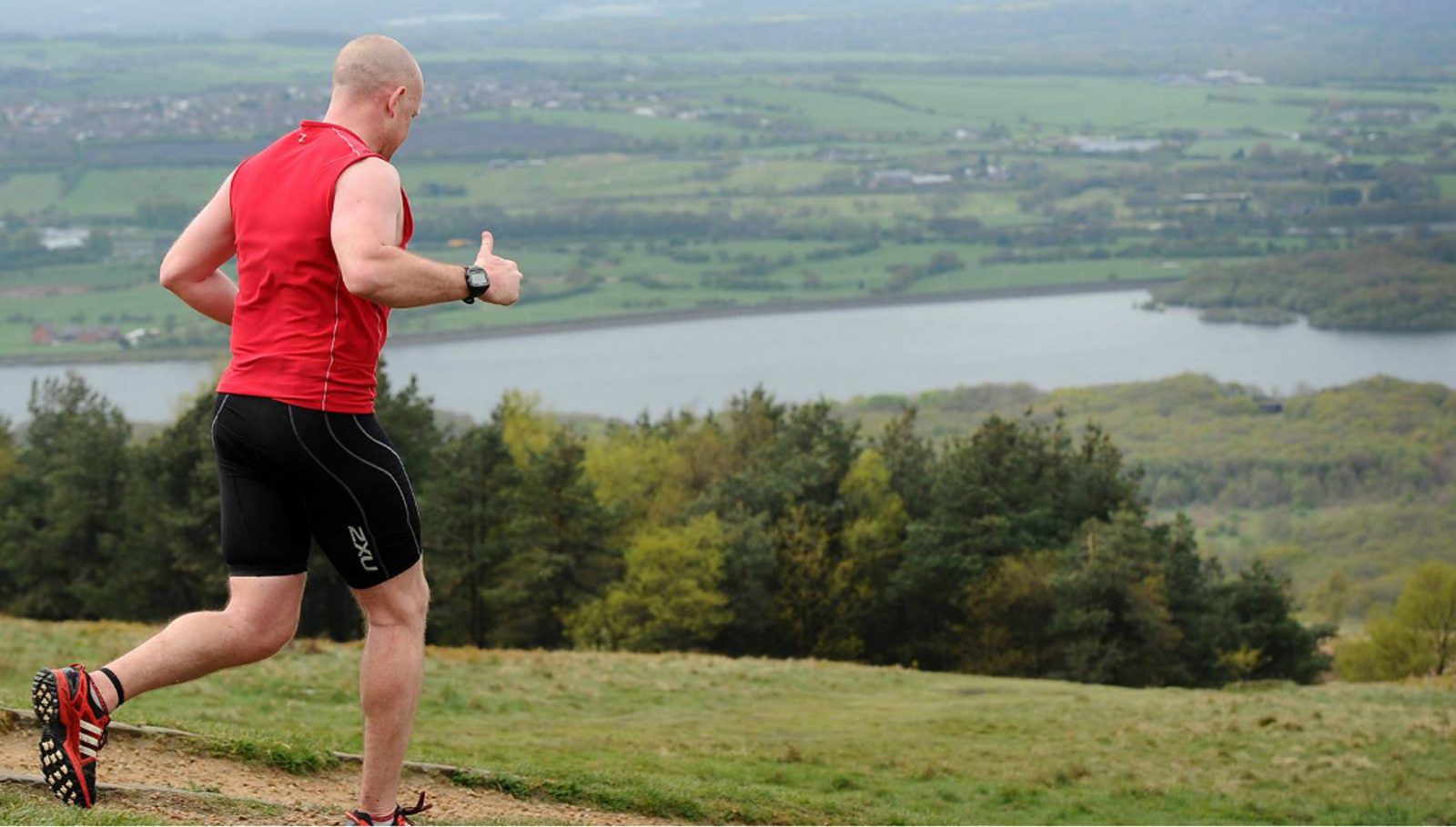 A man in a red sleeveless shirt and black shorts runs on a trail overlooking a scenic landscape with lush green trees, a river, and rolling hills under an overcast sky.