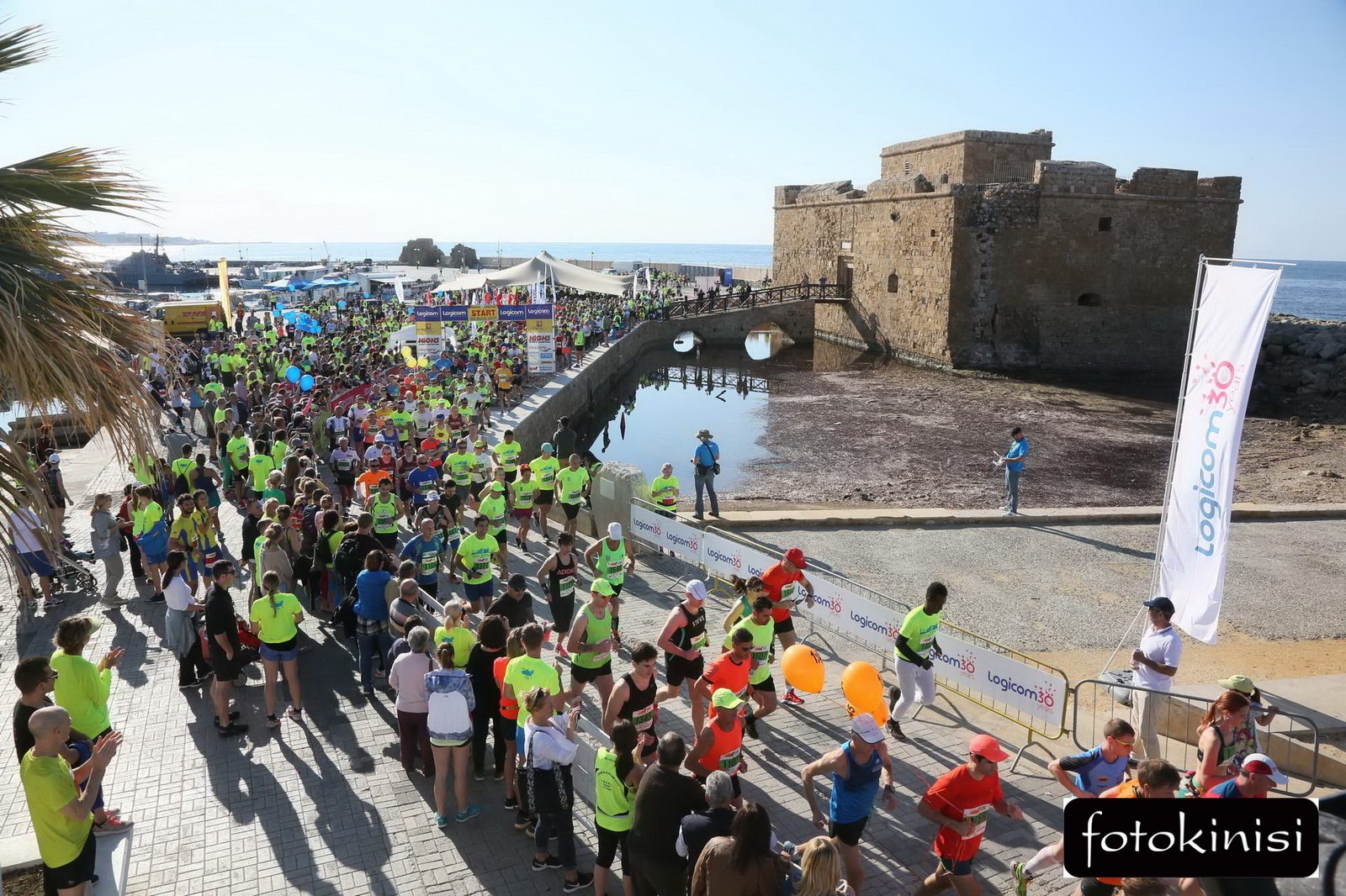A large crowd of people, many wearing bright green shirts, participate in an outdoor running event near a historic stone fort by the sea. The event features colorful balloons and banners, with spectators lining the route. The sky is clear and sunny.