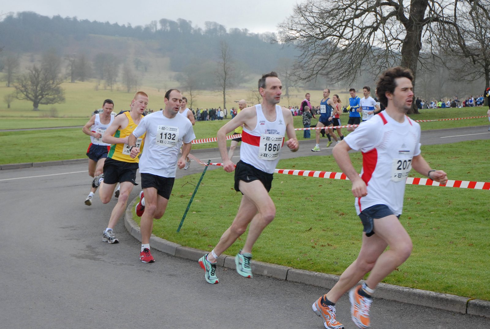 A group of runners in athletic gear are competing in a race on a paved road. One runner in a white shirt is ahead, followed closely by others with numbers pinned to their shirts. The background features grassy fields, trees, and spectators standing behind a barrier.