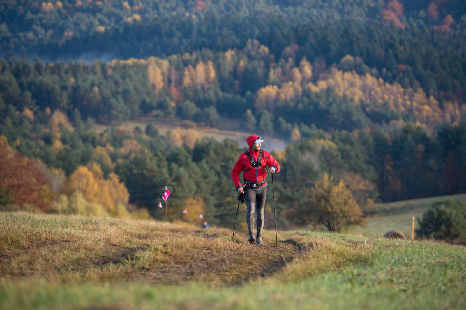 A person in a red jacket and black pants is hiking with poles along a grassy trail on a hillside. The background features a forested landscape with trees displaying autumn colors.