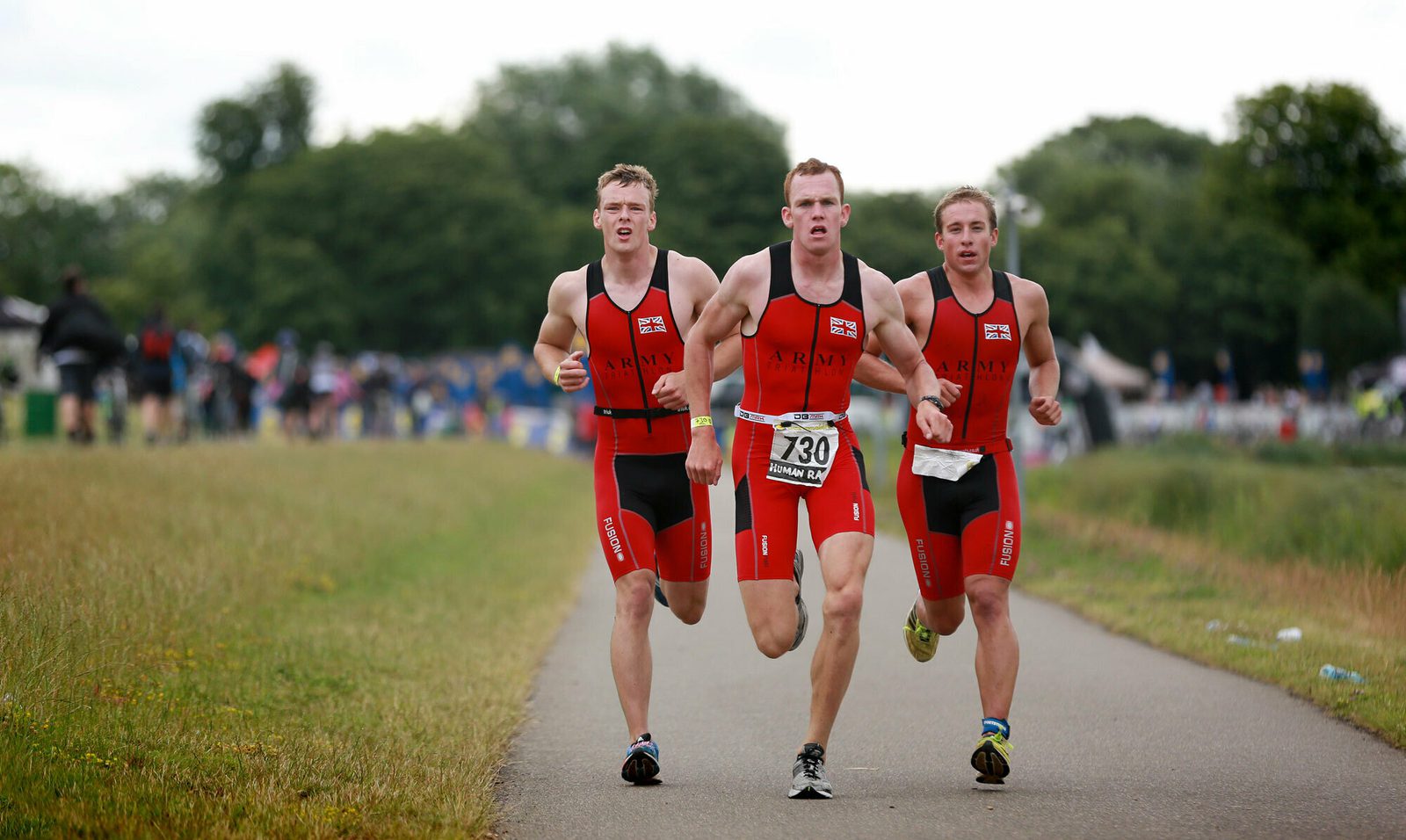 Three athletes wearing red triathlon suits with "ARMY" written on them are running on a paved path in a park-like area, with spectators in the background. The athlete in the center is wearing a bib number 730. It's a cloudy day with green trees surrounding the path.