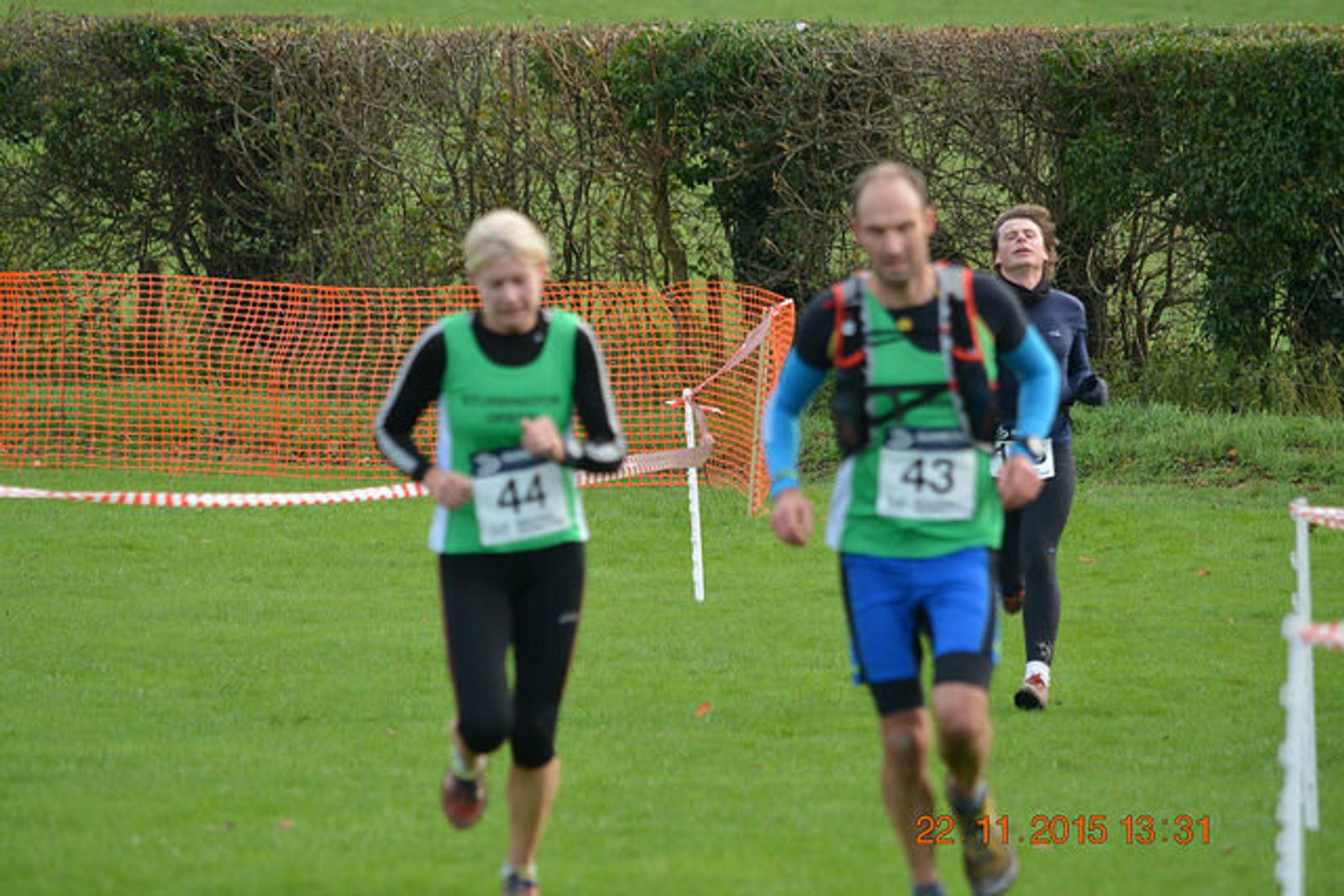 Three runners participating in a race are captured mid-action on a grassy field. The runner in the foreground is wearing a green top and black pants, while the others are similarly dressed in athletic wear. An orange mesh barrier and green foliage are in the background.
