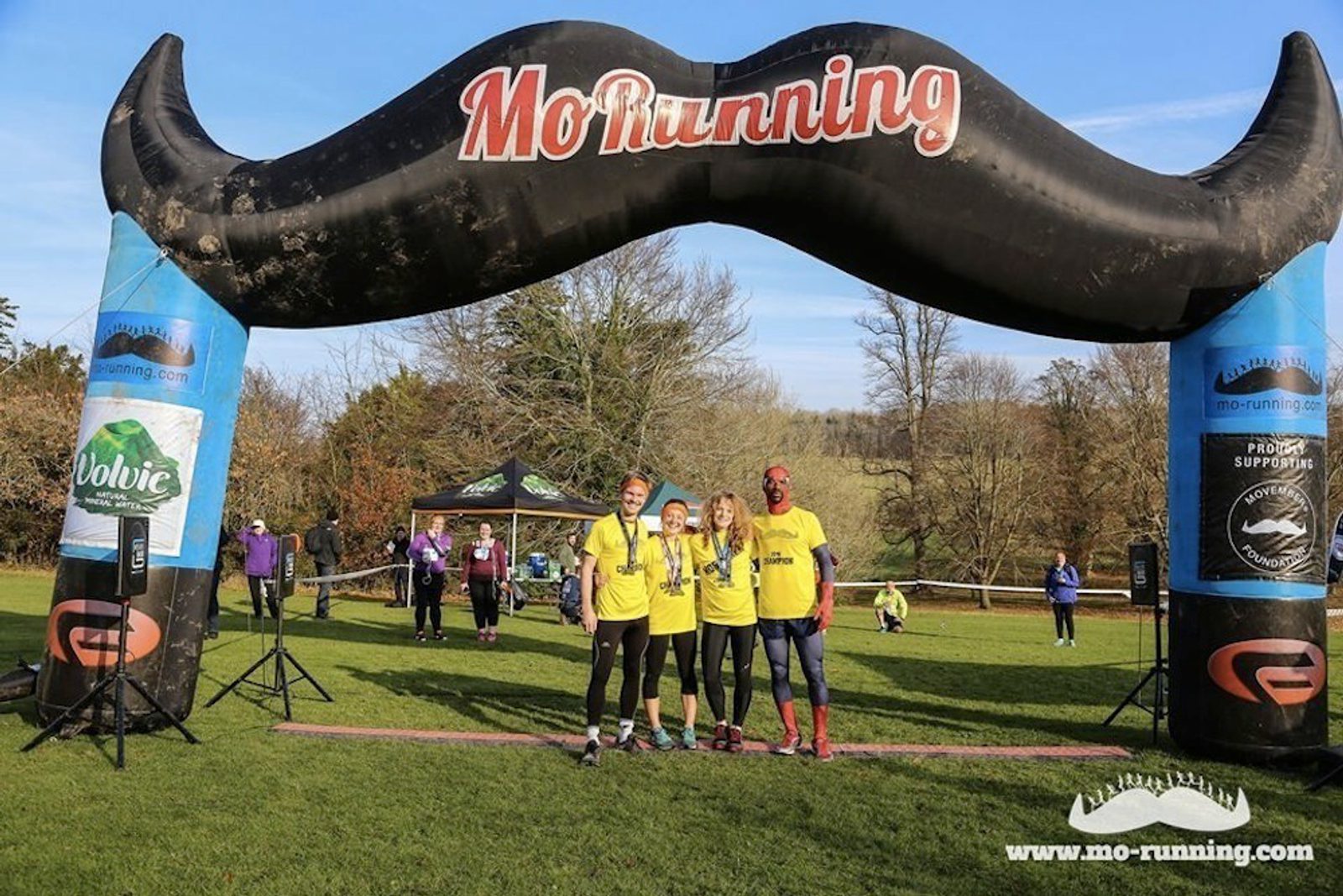 A group of four people in yellow shirts stand under a large inflatable mustache arch with "MoRunning" written on it. They are on a grassy field, smiling and holding medals. Other participants and event banners are visible in the background.