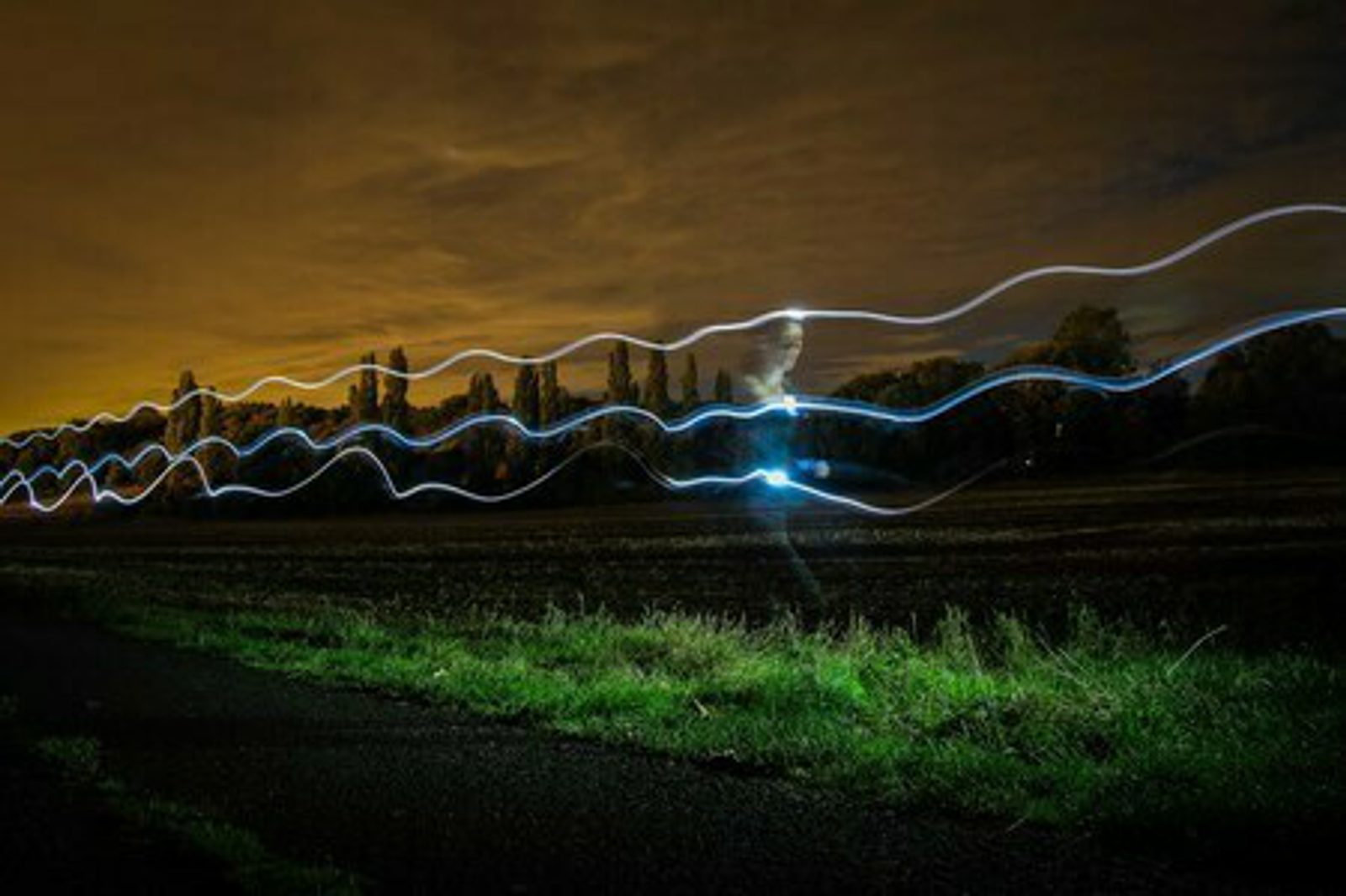 A nighttime scene featuring a field under a cloudy sky with tall trees in the background. Bright, wavy lines of light in blue and white streak across the sky and field, creating light trails that contrast with the dark surroundings. The foreground includes a grassy pathway.