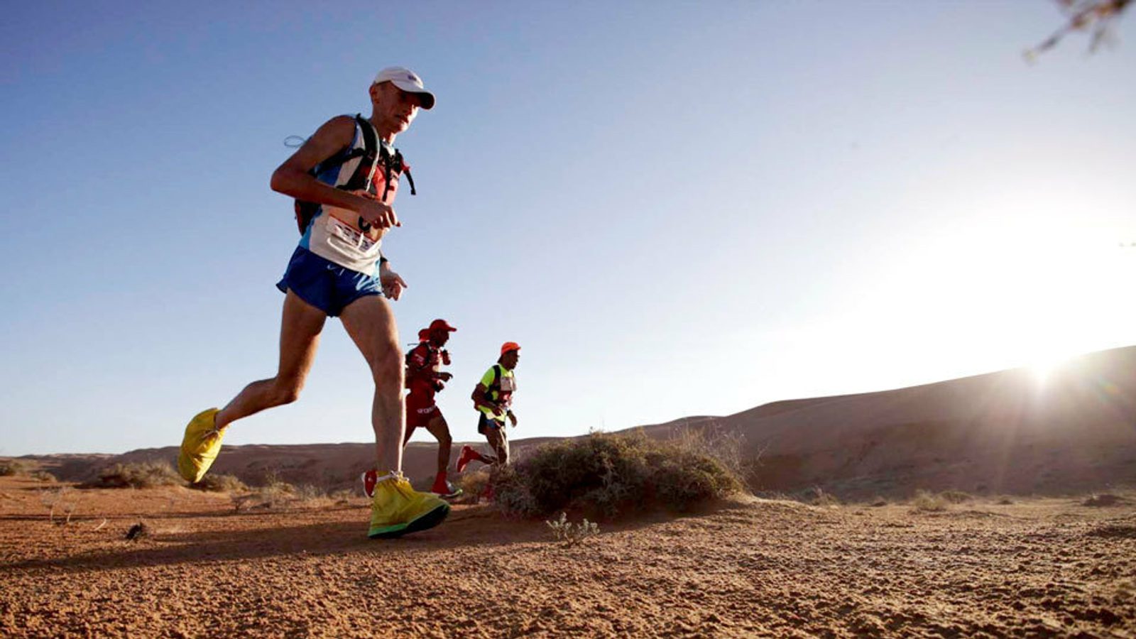 Several runners are participating in an outdoor race, traversing a barren desert landscape under a clear blue sky. One runner in the foreground is wearing a white hat, blue shorts, and yellow shoes, followed by two runners in colorful gear in the distance.