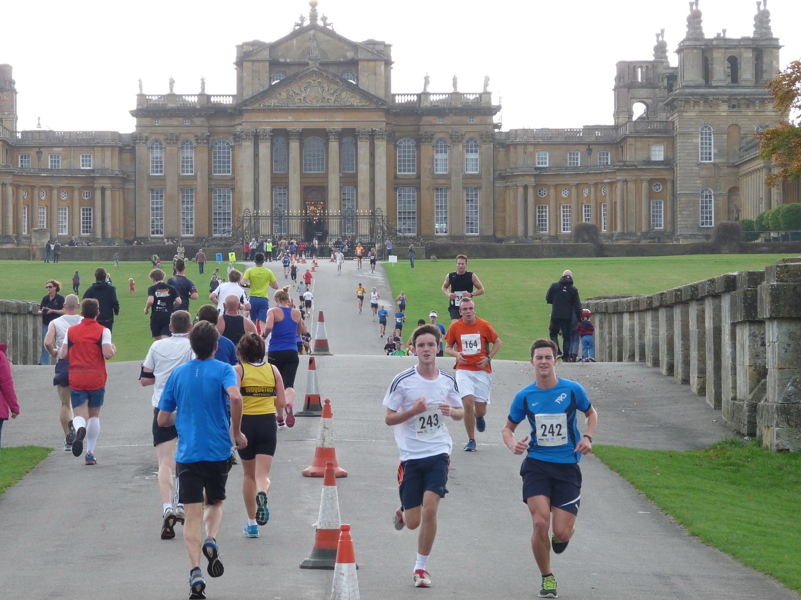 Participants in a race run along a pathway leading to a large historic building in the background. Some runners are heading toward the building, while others run in the opposite direction. The area is lined with traffic cones, dividing the pathway.