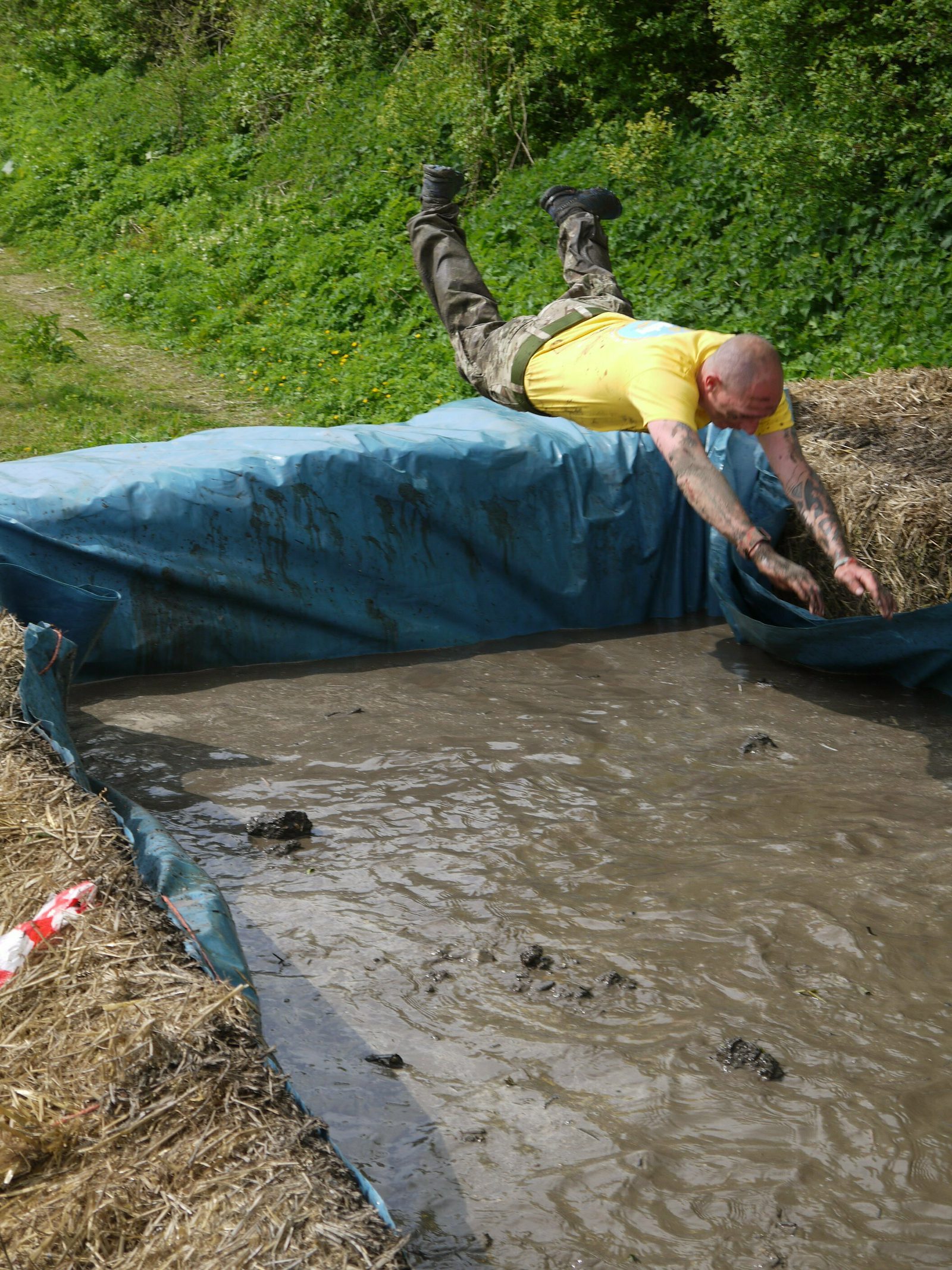 A man in a yellow shirt and camouflage pants dives headfirst into a mud pit during an outdoor obstacle course. The surrounding area is grassy and green. There are bales of hay on either side of the mud pit.
