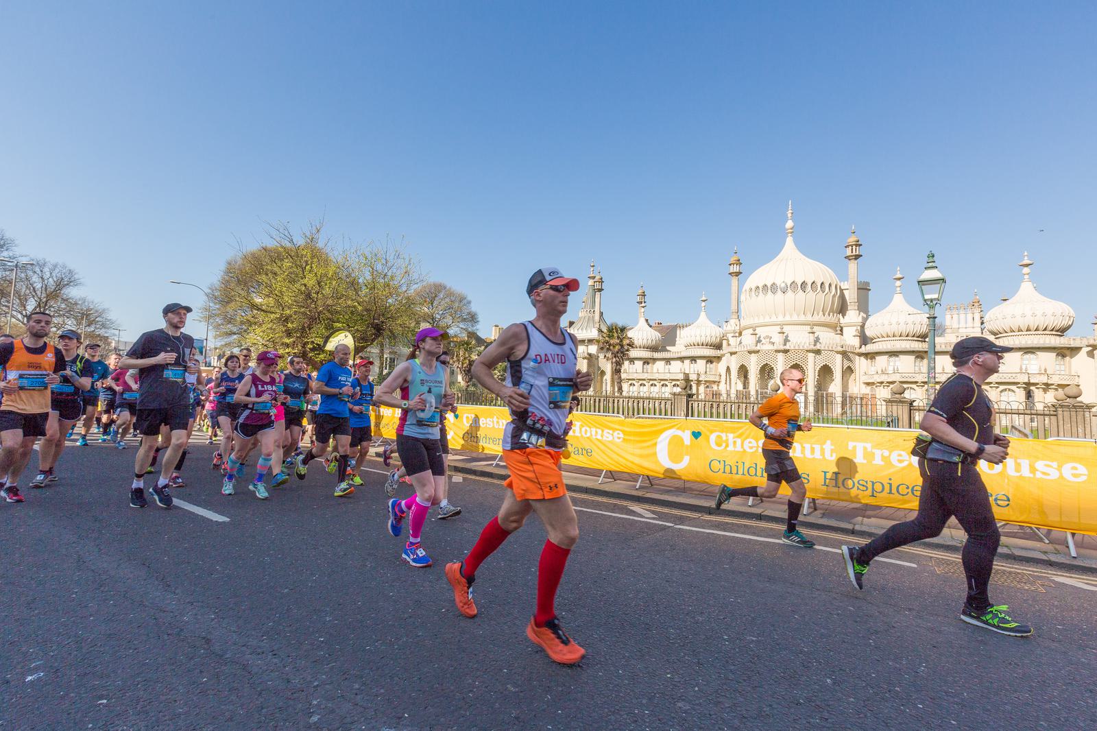 Runners compete in a marathon on a sunny day, passing by a historic, white palace with domes and spires. The runners are of various ages and wearing colorful athletic gear. A banner along the route advertises Chestnut Tree House Children's Hospice Care.