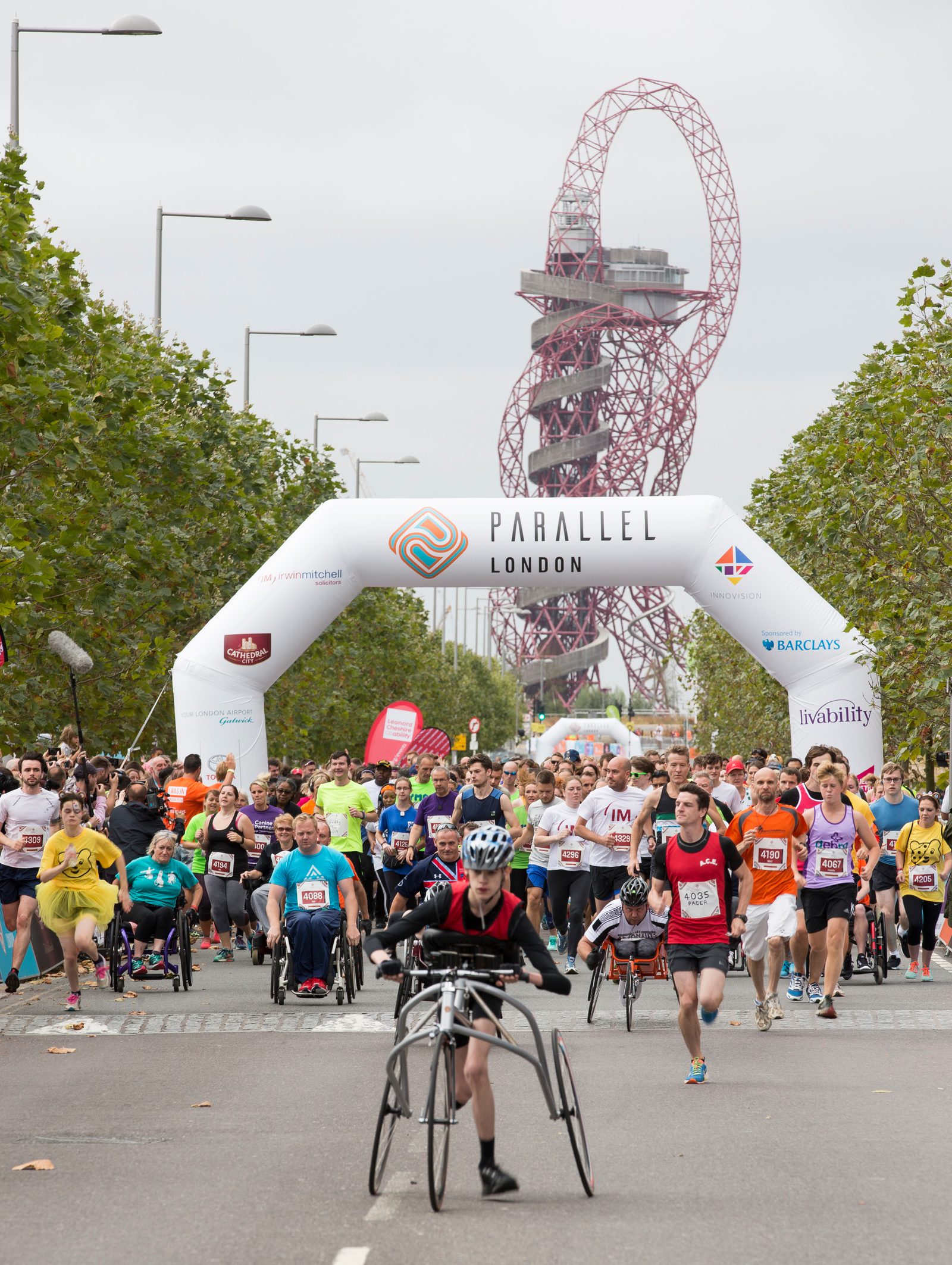 A diverse group of participants, including runners and individuals using wheelchairs, is seen starting the Parallel London race under an inflatable archway. The ArcelorMittal Orbit sculpture is visible in the background, framed by trees lining the road.