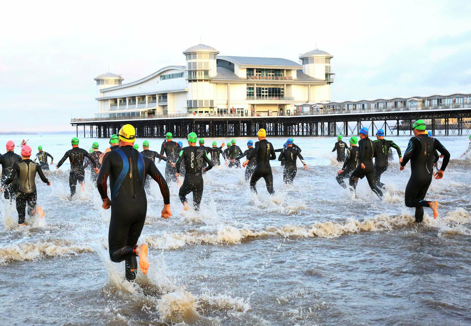 A group of triathletes in wetsuits and swim caps run into the ocean at the start of a race. The athletes are seen from behind, and in the background is a large pier with a multi-story building. The water is splashing around their legs as they enter.