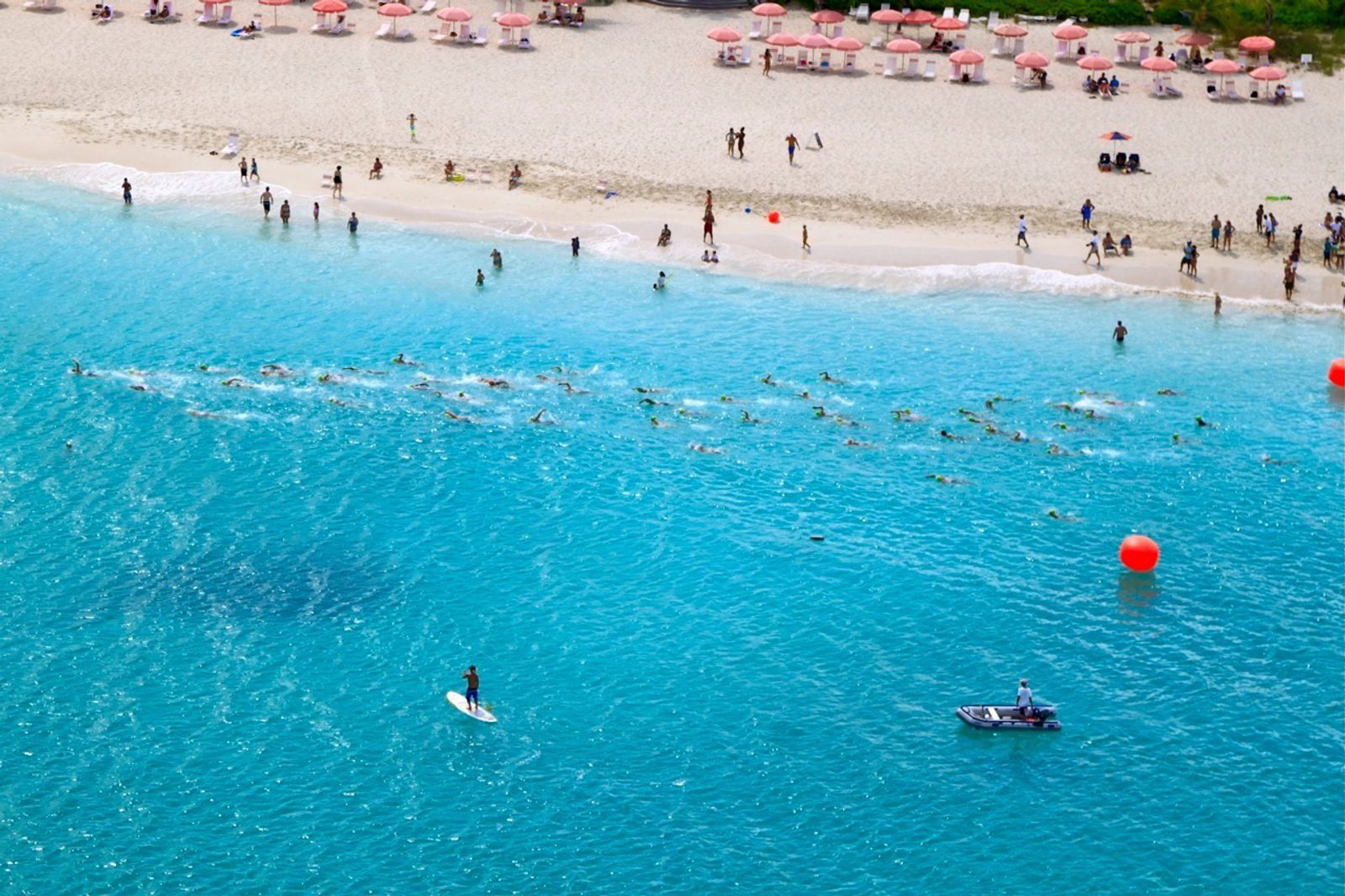 Aerial view of a beach scene with swimmers participating in an open water race marked by orange buoys. People relax under pink umbrellas on the sandy shore. A paddleboarder and a small boat are nearby in the turquoise water.