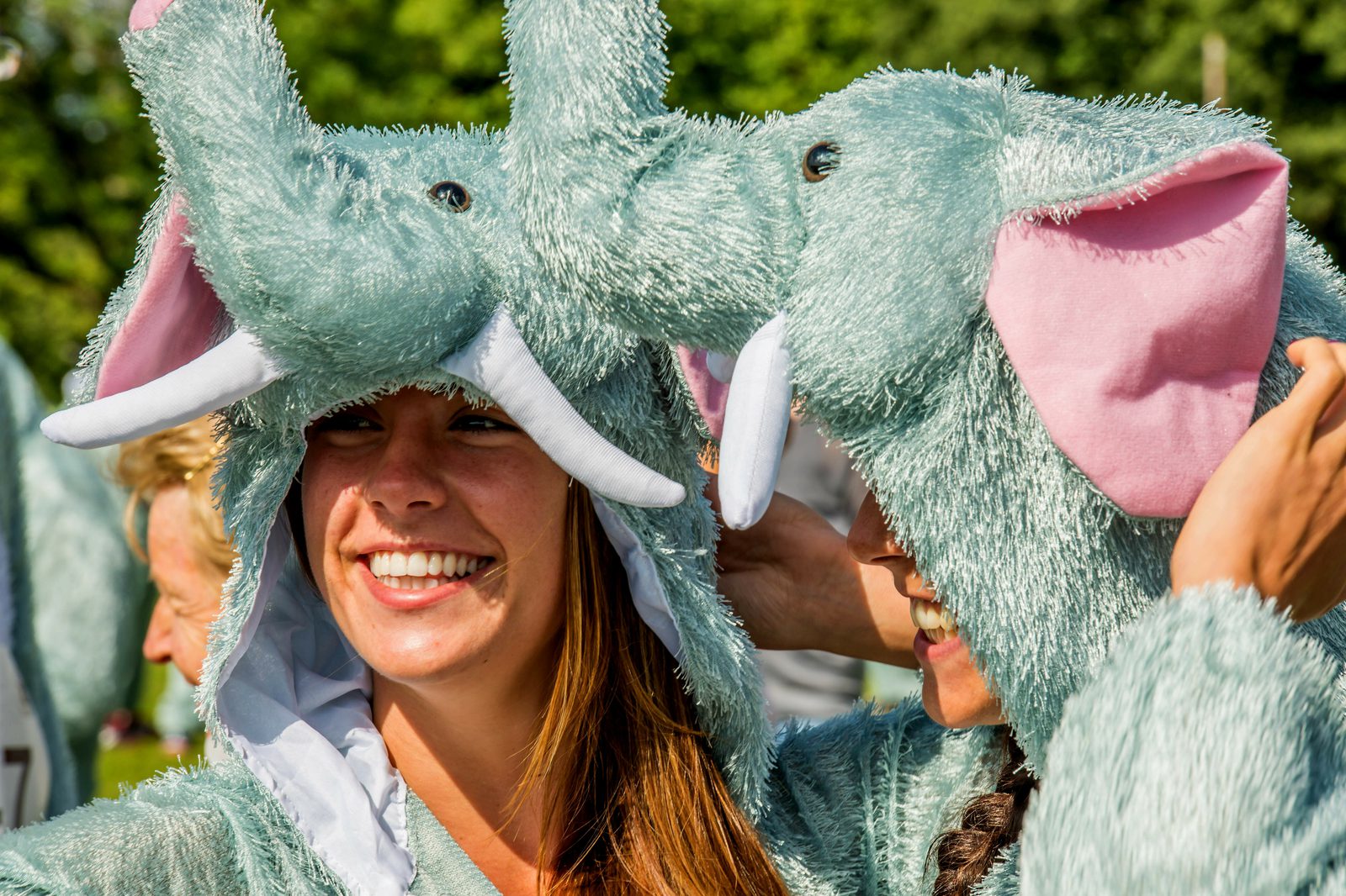 Two people wearing fluffy elephant costumes with long trunks and pink inner ears smile joyfully. They appear to be outdoors with greenery in the background, enjoying a festive or playful event.