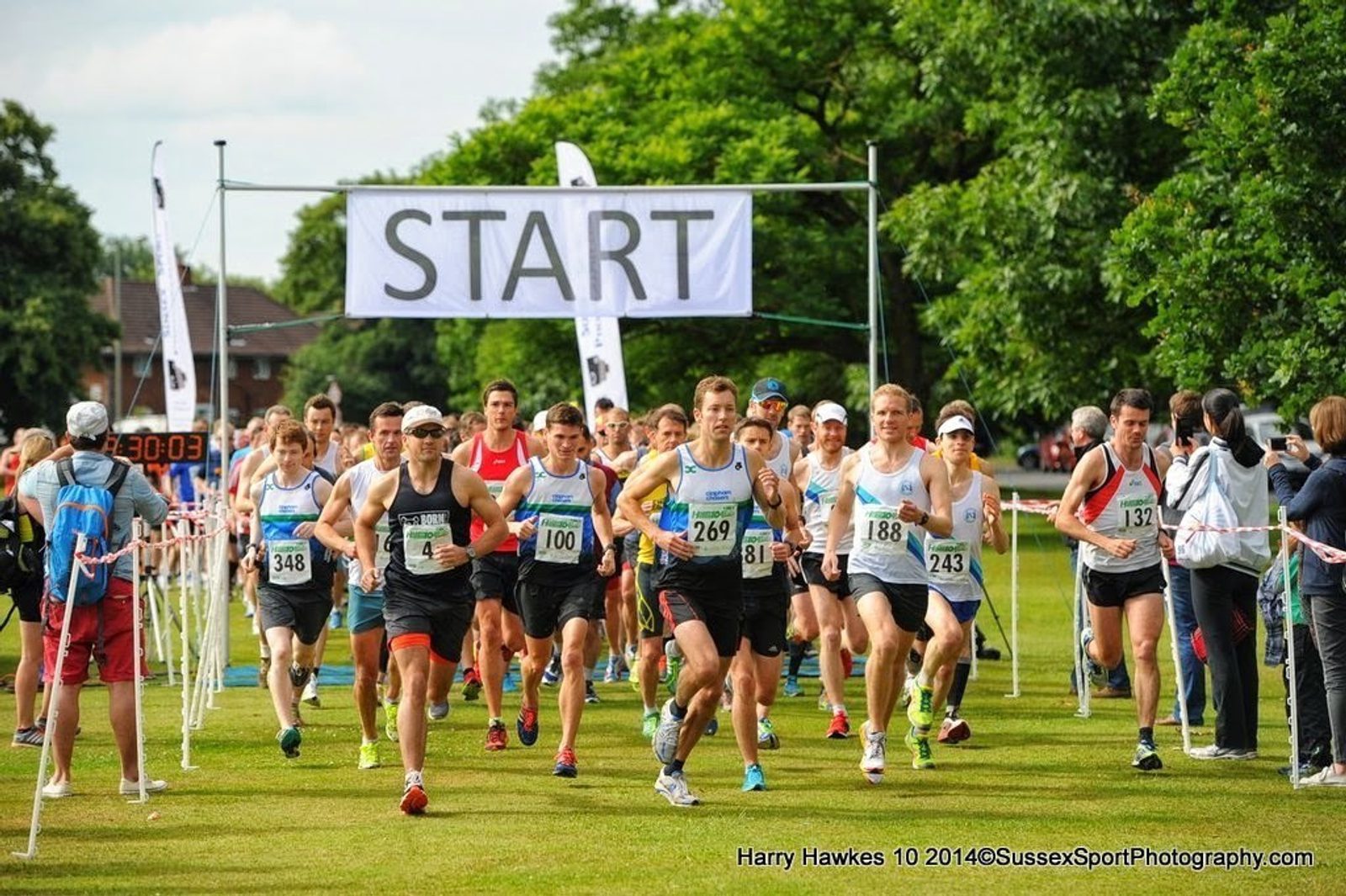 A group of runners begin a race, starting under a large banner that reads "START." They are running on a grassy field, surrounded by green trees and spectators. The runners are wearing race bibs with numbers, and the atmosphere appears vibrant and energetic.