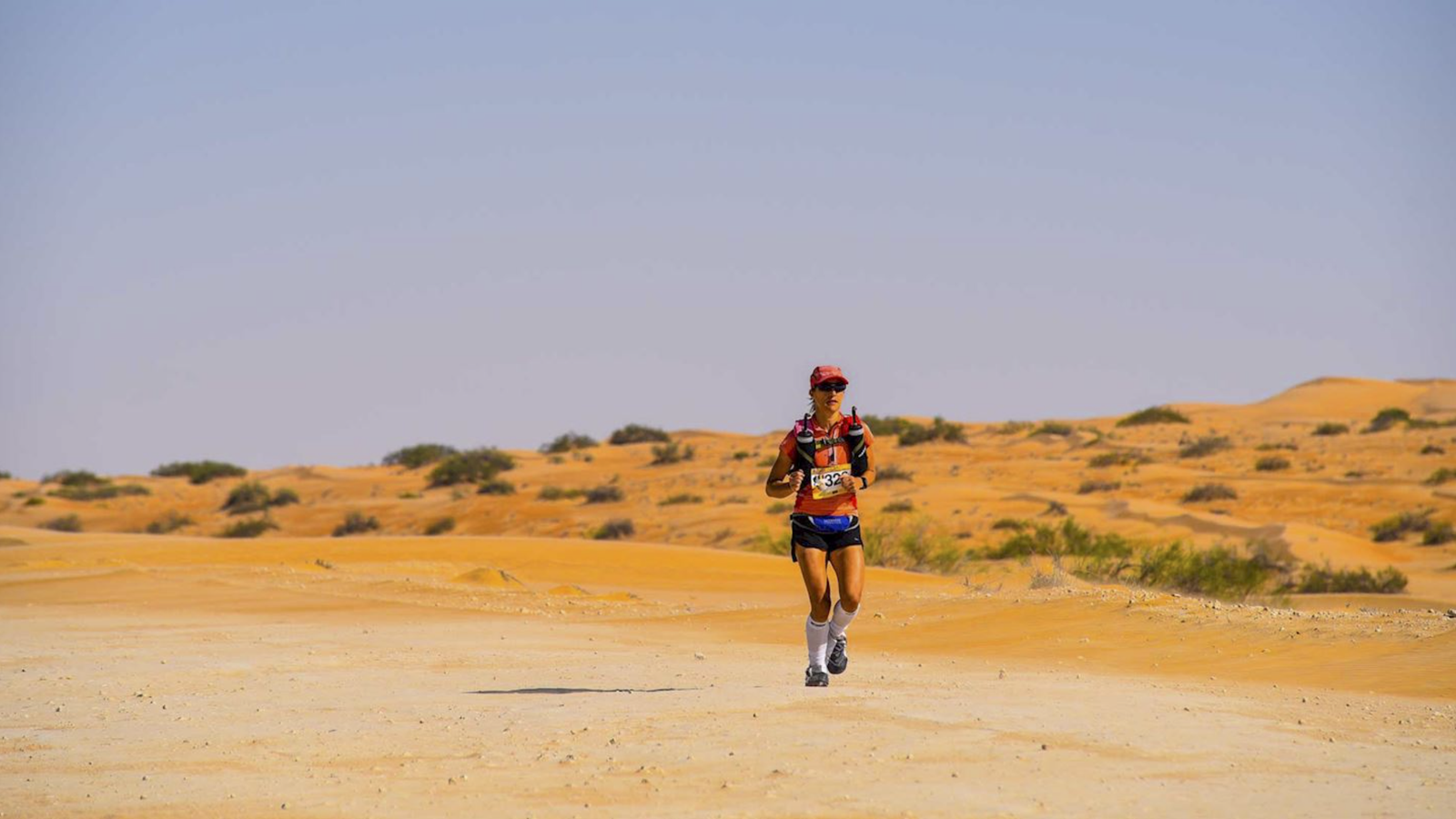A runner wearing a red cap, sunglasses, shorts, and a hydration pack jogs across a desert landscape with sparse vegetation under a clear blue sky. The ground is sandy and appears expansive, stretching out towards distant sand dunes.