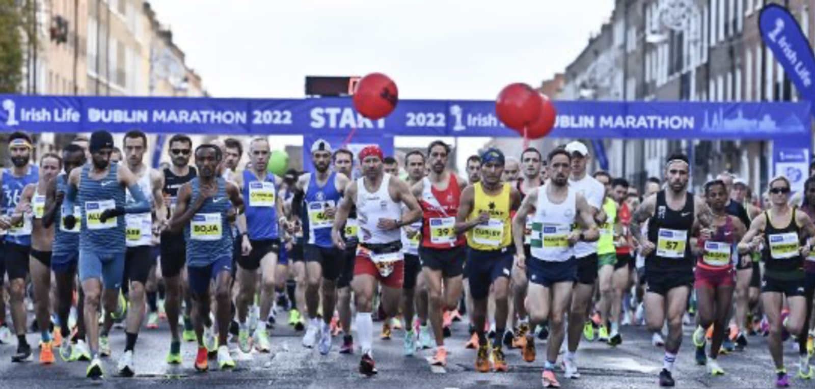 A group of runners, wearing various athletic outfits, begin the Dublin Marathon 2022. The starting line features a banner that reads "Irish Life Dublin Marathon 2022." Some runners hold red balloons, and the race takes place on a city street lined with buildings.