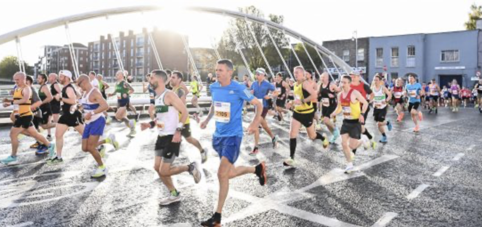 A large group of runners participates in a road race, captured mid-stride on a city street. The background features a modern white arch bridge and urban buildings. The runners wear diverse athletic gear and appear focused. Sunlight casts a bright glow on the scene.