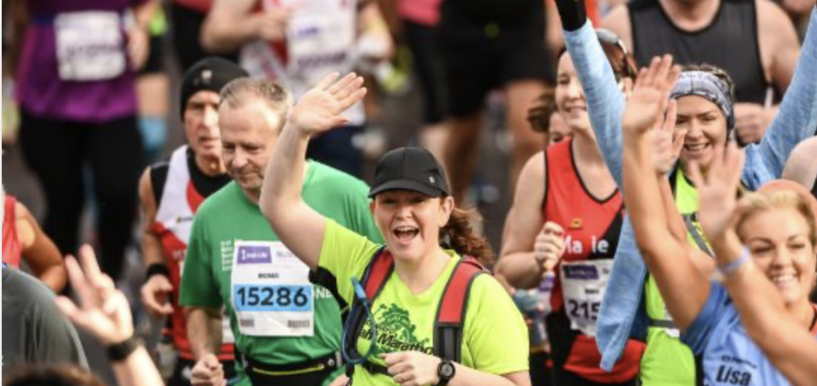 A group of runners participating in a race, wearing colorful outfits and bib numbers. The central figure, a smiling woman in a neon green shirt and black cap, waves enthusiastically. Other runners around her exhibit a mix of determination and joy.