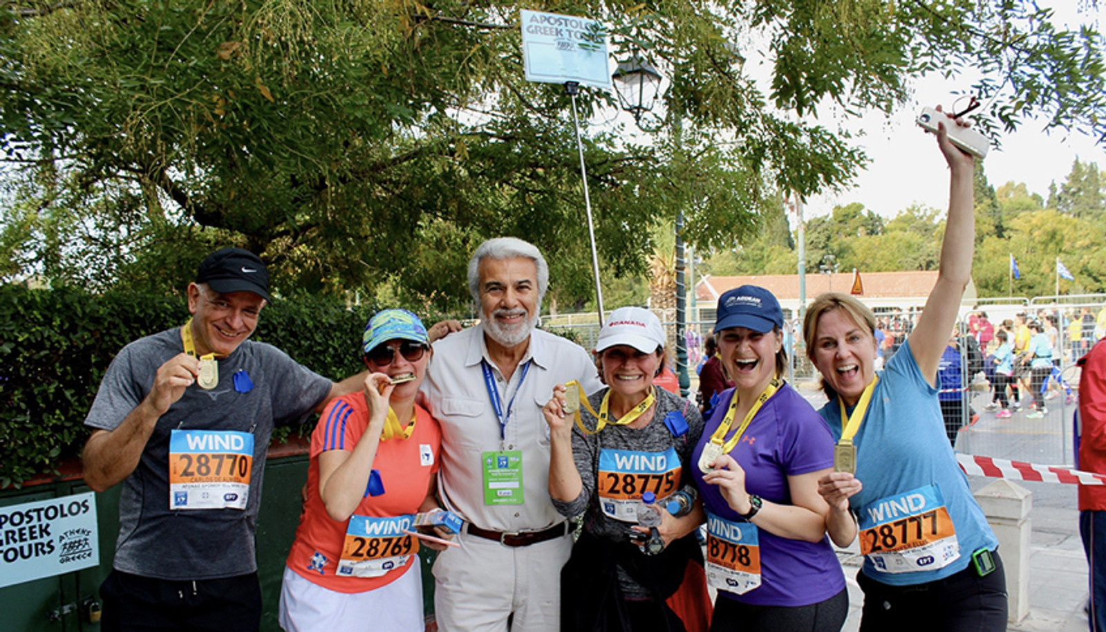 A group of six people, some wearing athletic gear and race bibs, celebrate together at the finish line of a race. They are smiling and holding up medals. Trees and a "Apostolos Greek Tours" sign are visible in the background.