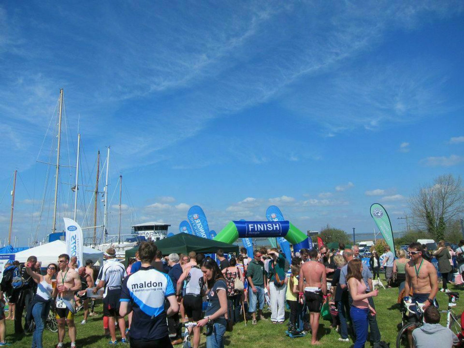 A large crowd gathers outdoors near a finish line arch at a sporting event. Sailboats and blue sponsor flags are seen in the background. Participants, some wearing sports attire and medals, and spectators enjoy the sunny day under a blue, cloud-streaked sky.