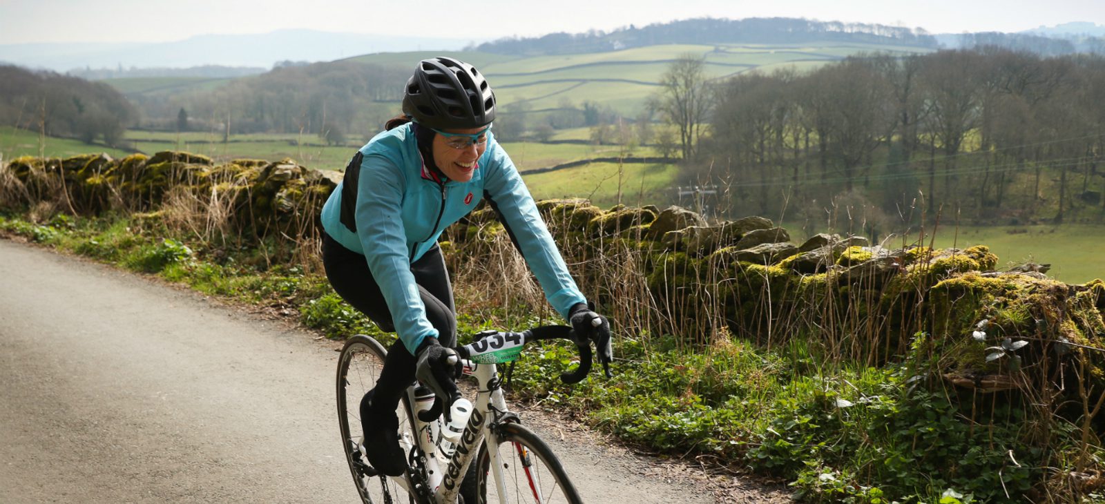 A cyclist wearing a blue jacket, black helmet, and glasses rides along a country road bordered by a moss-covered stone wall and scenic green fields. The rolling hills and leafless trees are visible in the background on an overcast day.