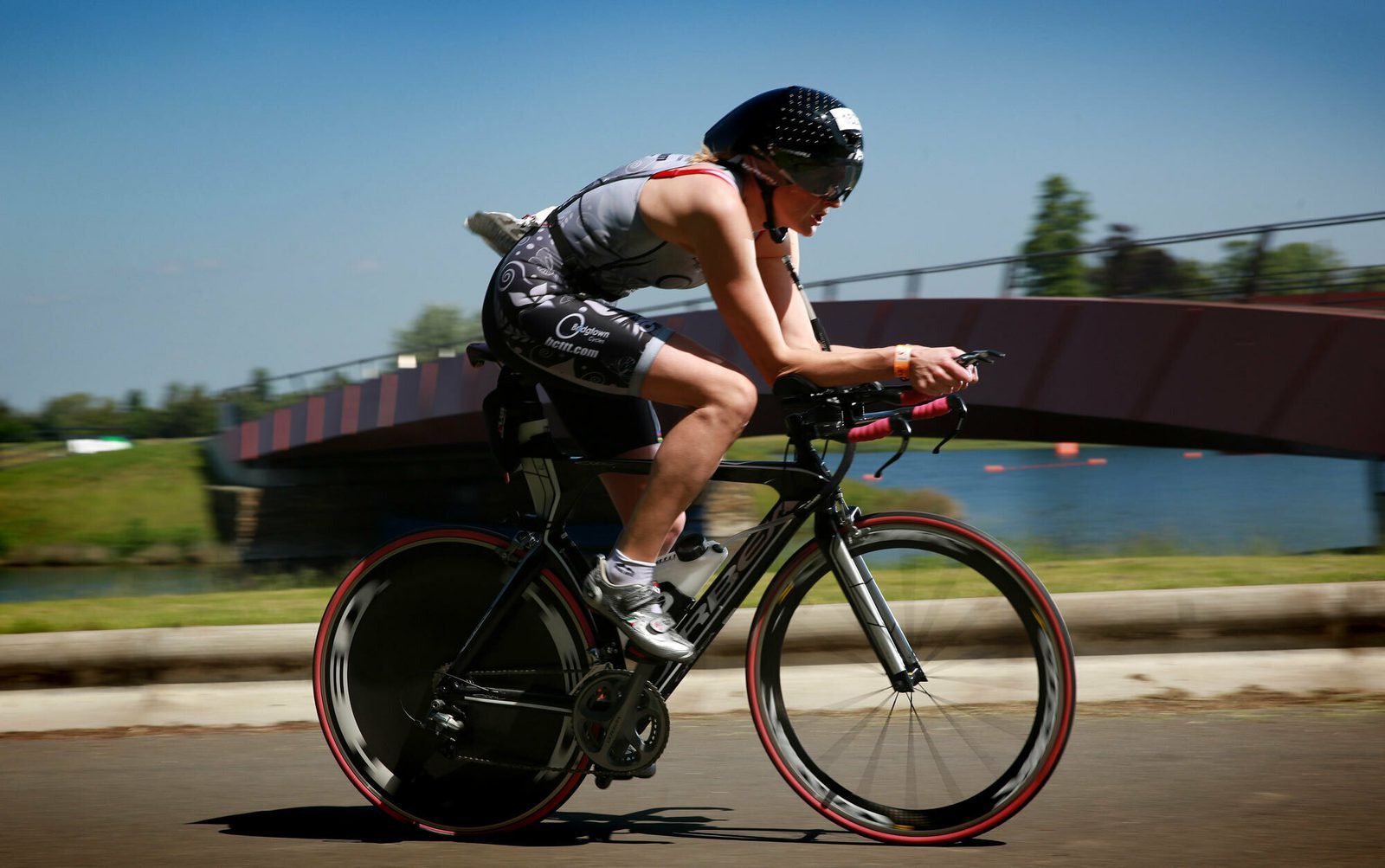 A cyclist competes in a race, wearing a helmet and aerodynamic gear. The rider is hunched over the handlebars, focusing on speed. They ride a high-performance bike with disc wheels, passing by a bridge. The background is a clear, sunny day near a body of water.