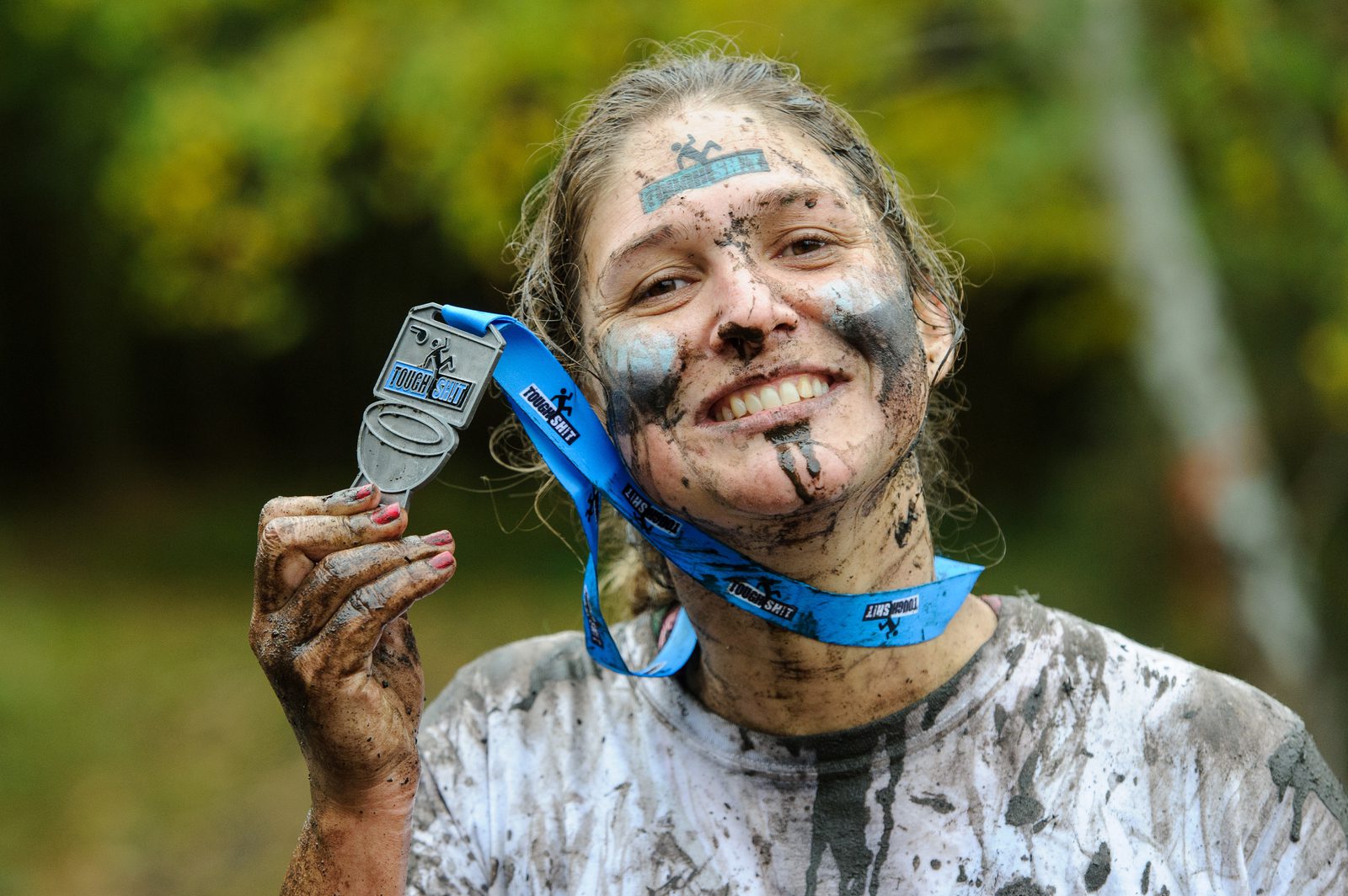 A mud-covered person smiles while holding up a medal by its blue ribbon. They have face paint and wear a white, mud-soaked shirt, suggesting they've just completed a challenging outdoor event. The background shows green, blurred foliage.