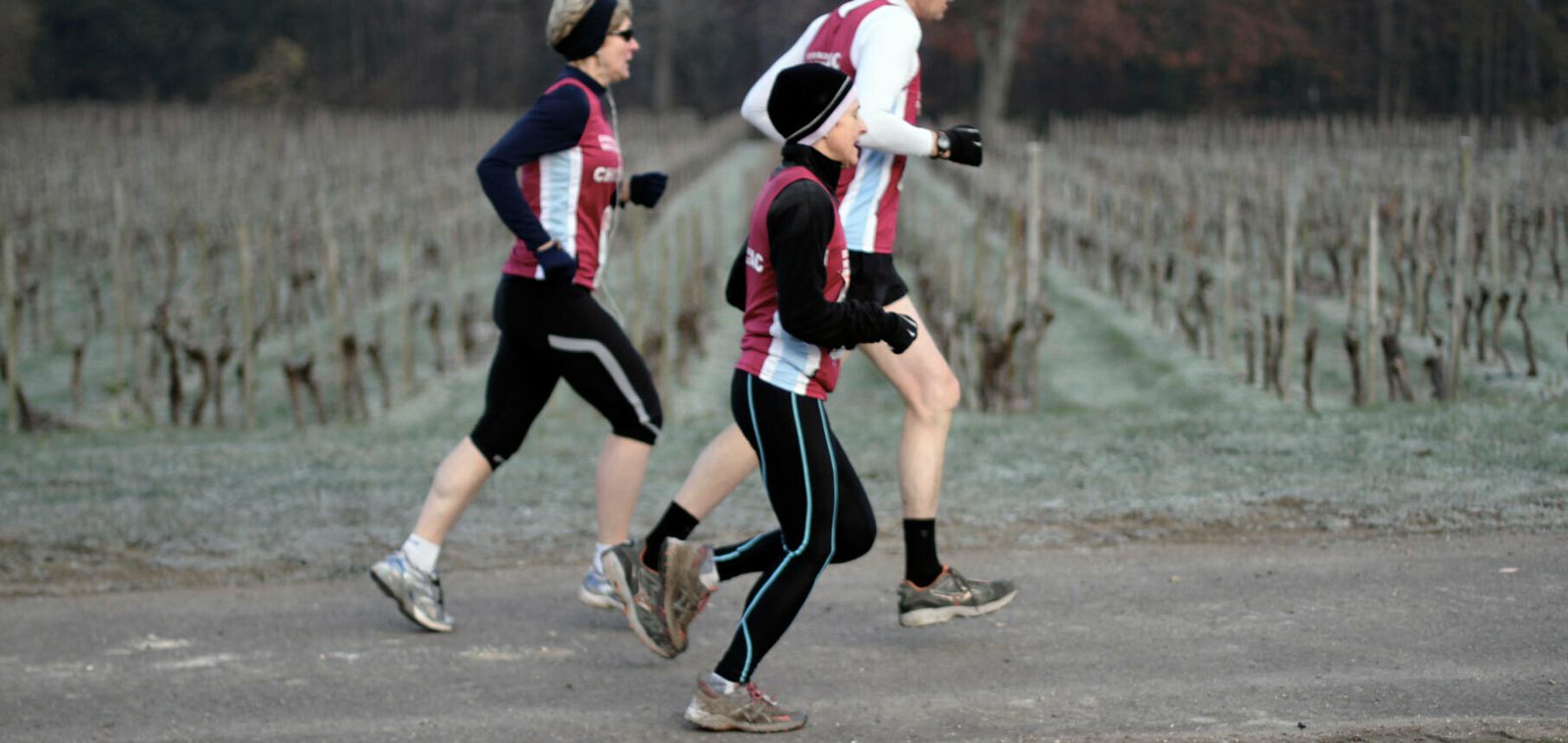 Three people jogging on a paved path in a vineyard or agricultural area. They are wearing athletic clothing, including matching red and white sleeveless tops, and are running in the same direction. The weather appears cool and the ground is slightly frosty.
