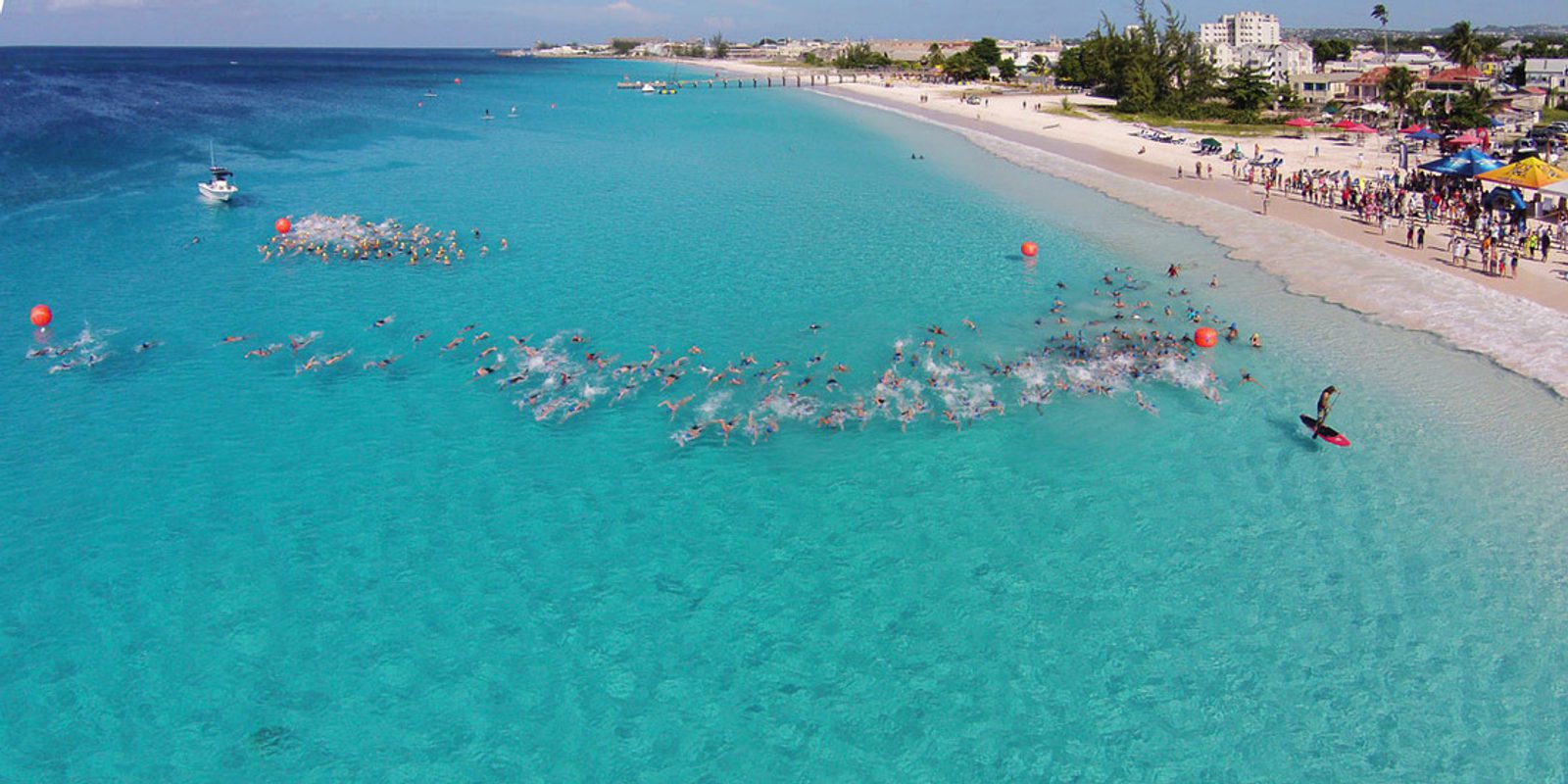Aerial view of a crowded beach with numerous swimmers participating in a large open water swimming event. The clear blue ocean stretches out to the horizon. Spectators are gathered along the shore, and several red buoys mark the swimming route.