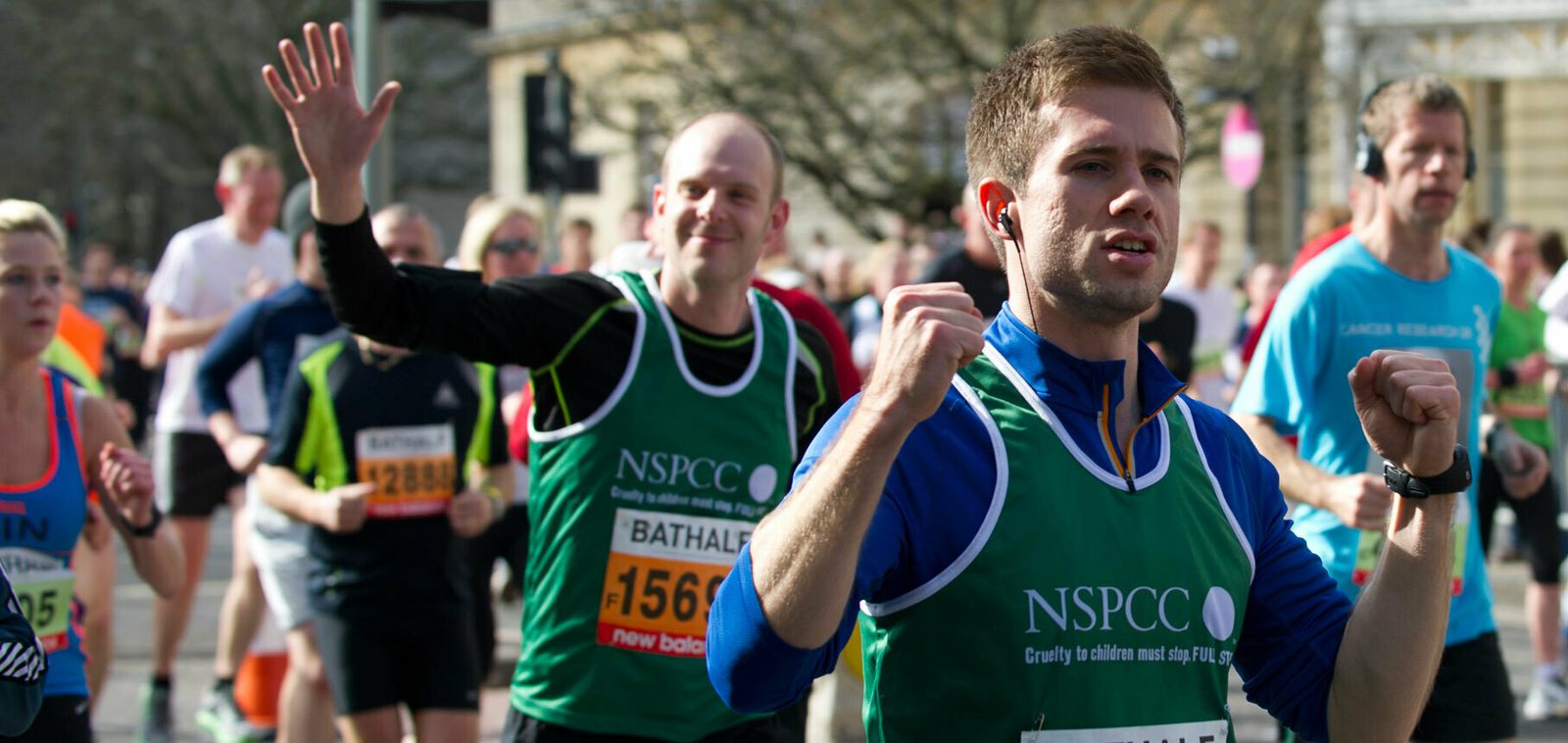Runners participating in a road race, with many wearing bright-colored athletic attire. Two runners in green NSPCC vests are in the foreground, one of whom is smiling and waving, while the other is running with headphones. The background shows other participants and trees.