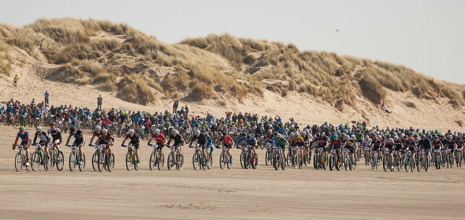 A large group of cyclists is participating in a race on a sandy beach, with grassy dunes in the background. Crowds of spectators watch from the dunes. The sky is clear, suggesting fair weather for the event.
