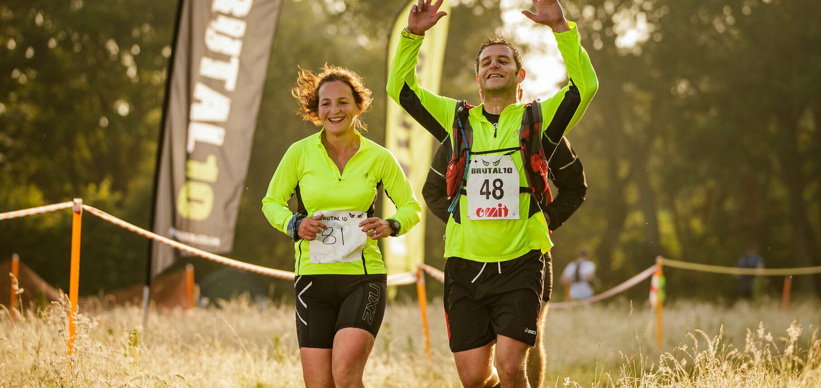 Two runners in neon yellow shirts are crossing a grassy finish line under sunlight, smiling and celebrating. One has a number 48 bib and both appear joyous and exhausted, with a banner visible on the side and blurred trees in the background.