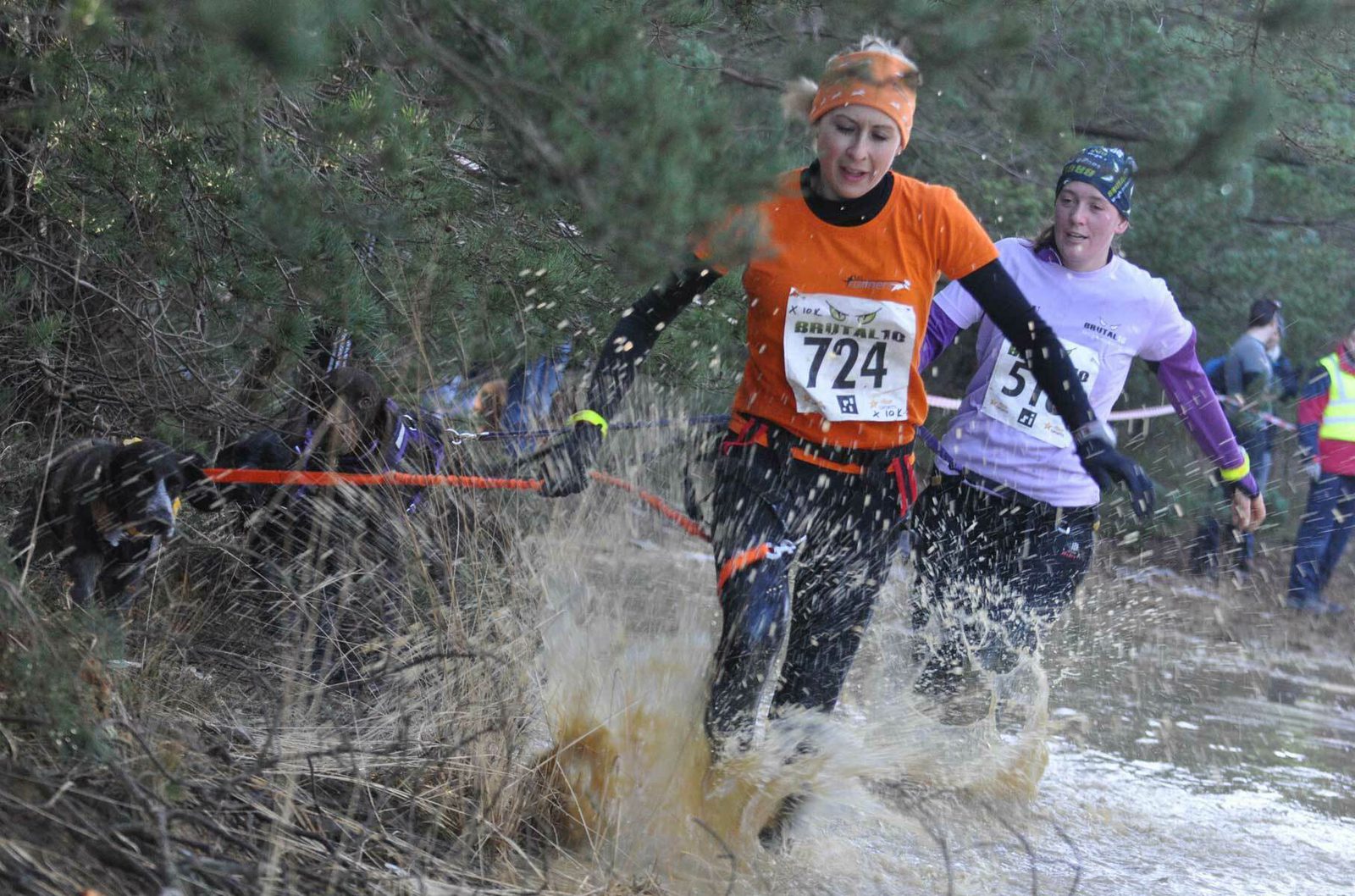 Two women compete in a muddy outdoor race. The woman in the foreground, wearing an orange shirt with the number 724, navigates through a shallow, muddy water puddle. The second woman, in a white shirt with the number 518, follows closely behind. Trees are in the background.