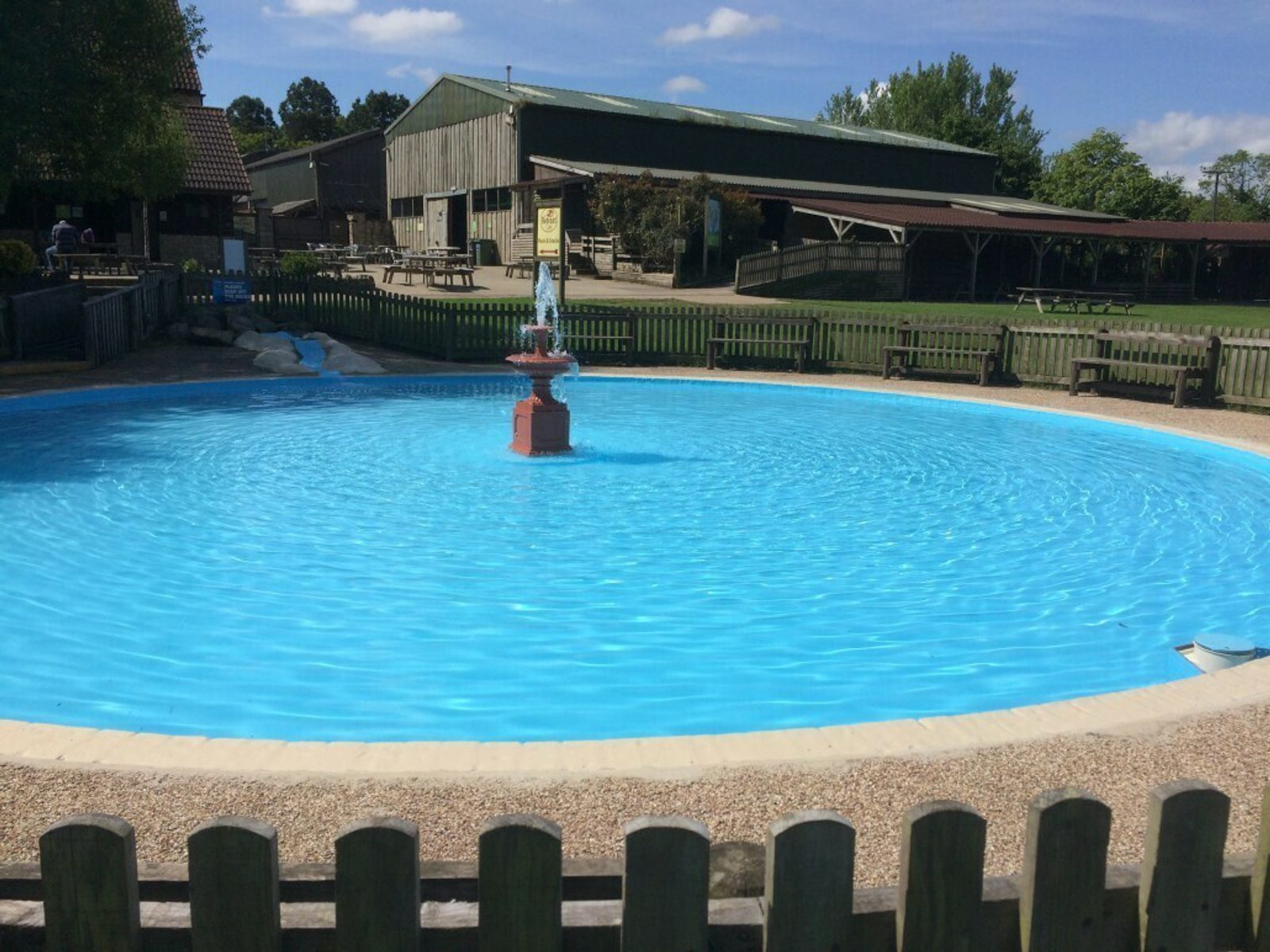 A circular water fountain in a blue pool is surrounded by a wooden fence. In the background, there are wooden buildings and picnic tables under a sunny, blue sky with scattered clouds. Benches are placed near the pool, and trees are visible in the distance.