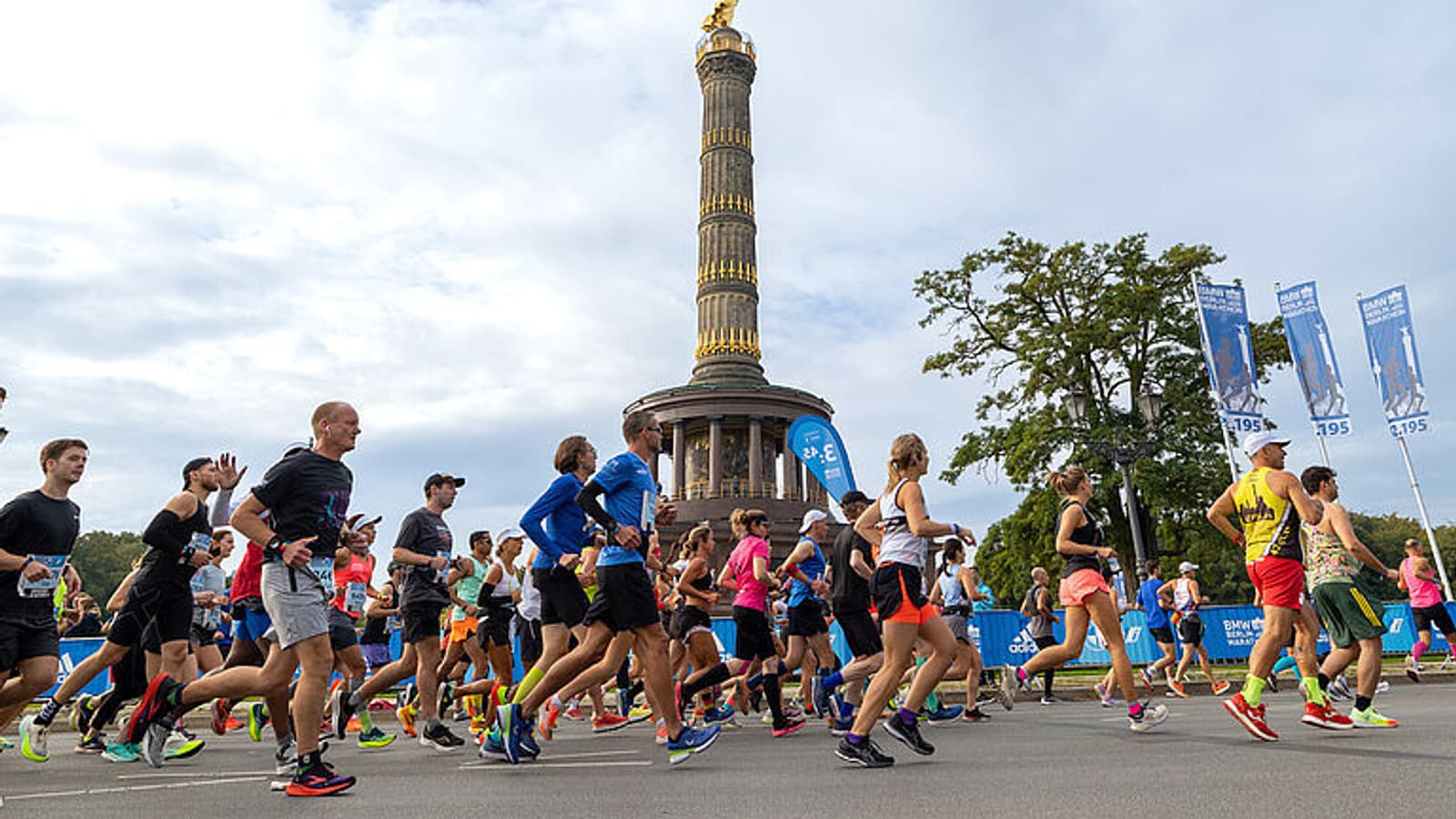 Participants run in a marathon passing by the Berlin Victory Column on a cloudy day. The runners are wearing various athletic outfits, while spectators and trees are visible in the background. Several blue banners are displayed along the route.
